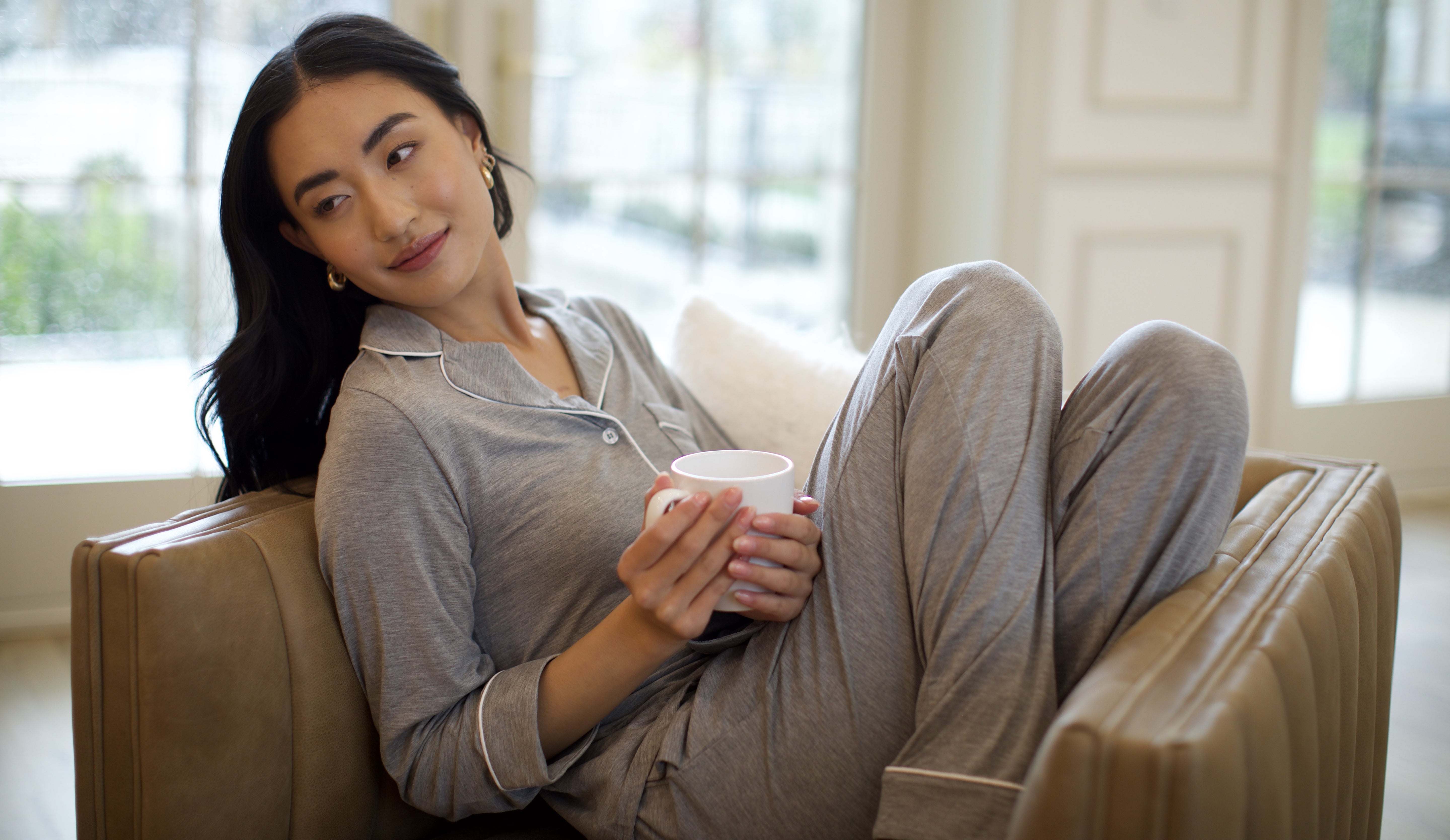 A person with long dark hair, wearing gray pajamas, is sitting on a brown couch holding a white mug. They are looking to the side, appearing relaxed in a cozy, well-lit room with large windows.