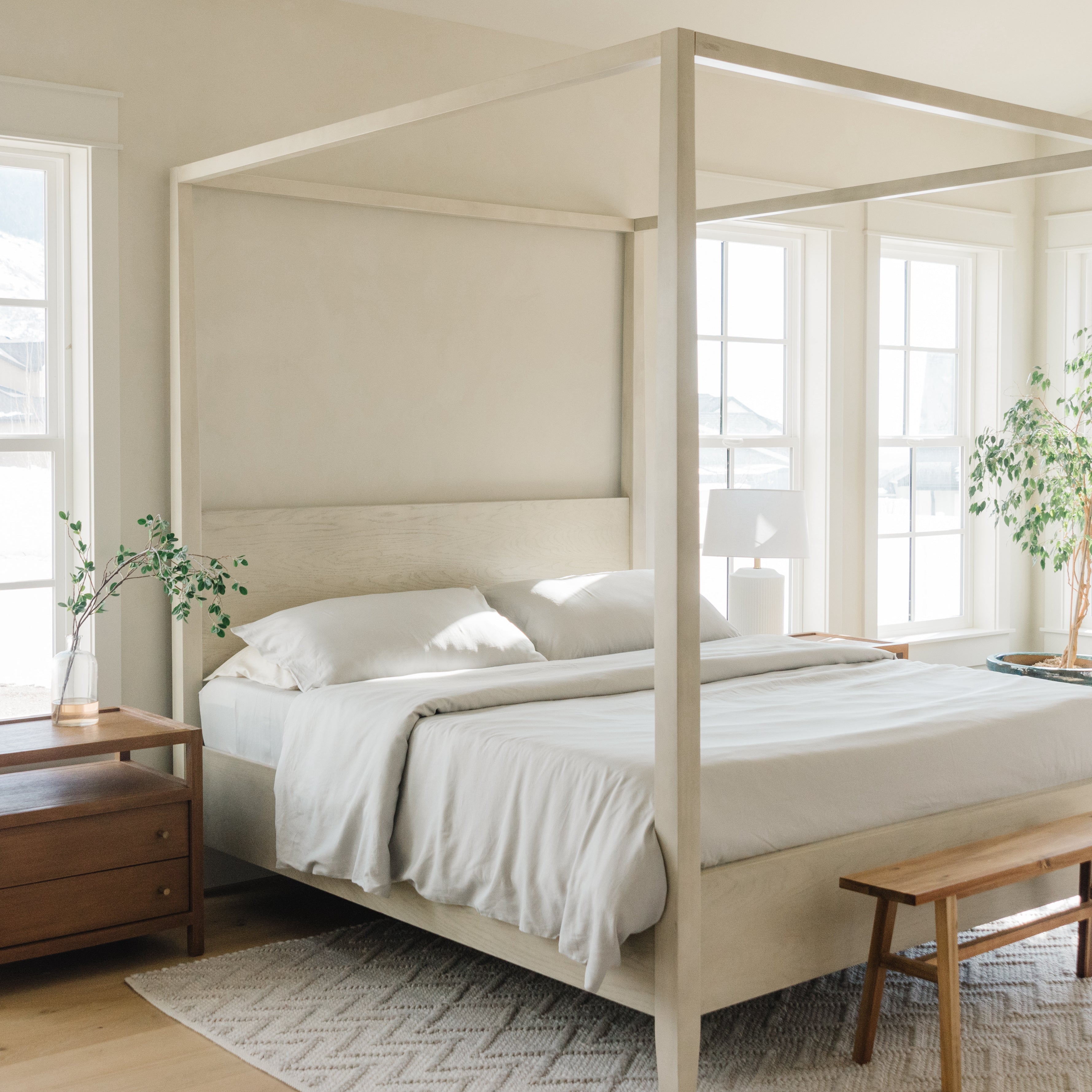 Bright, minimalist bedroom with a light wood canopy bed, white bedding, wooden bench, nightstand, lamp, indoor plants, and large windows letting in natural light.