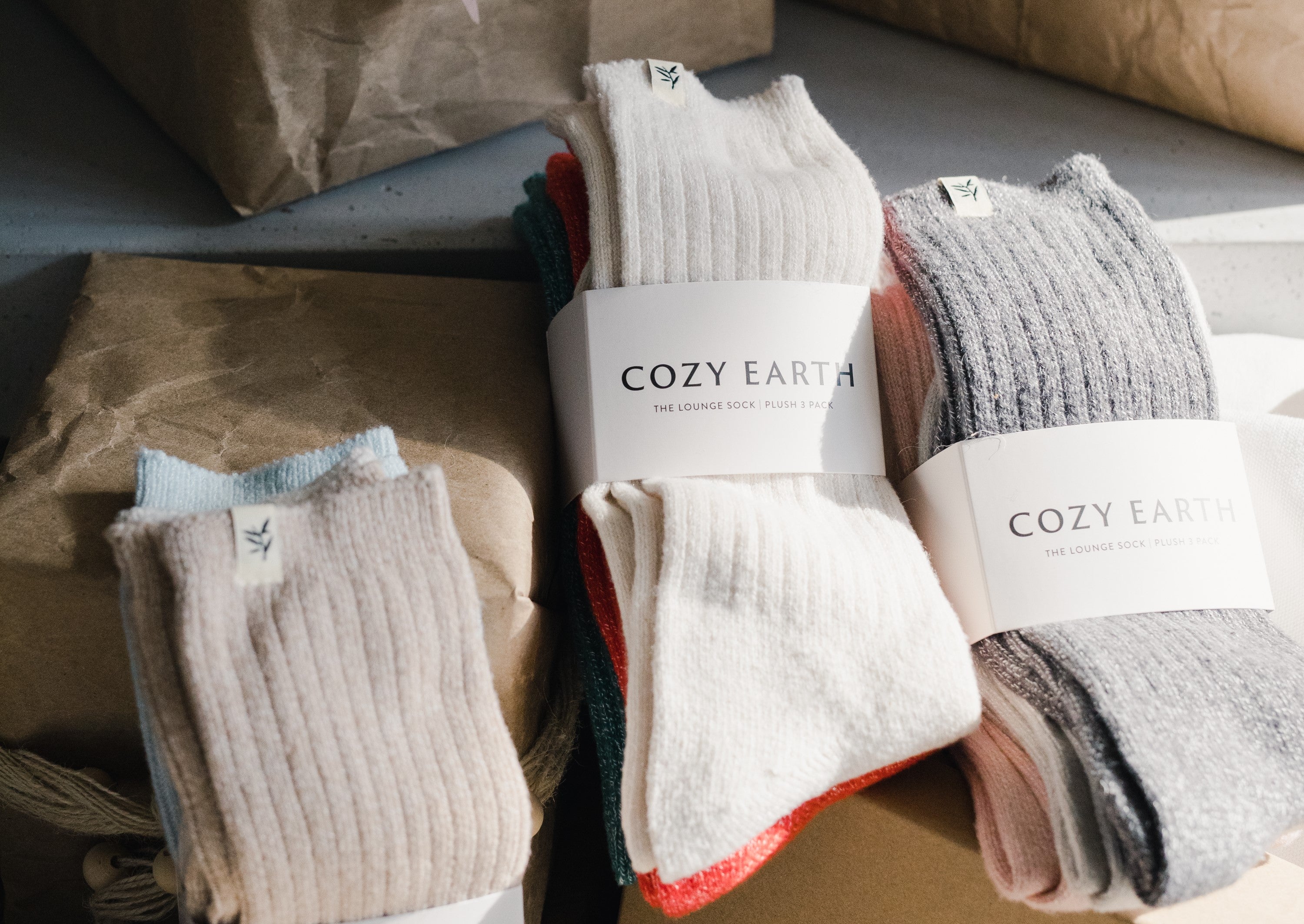 Three pairs of Cozy Earth socks, in beige, white, and gray, are displayed with their labels. They are placed on brown paper packaging, suggesting a cozy and eco-friendly presentation.