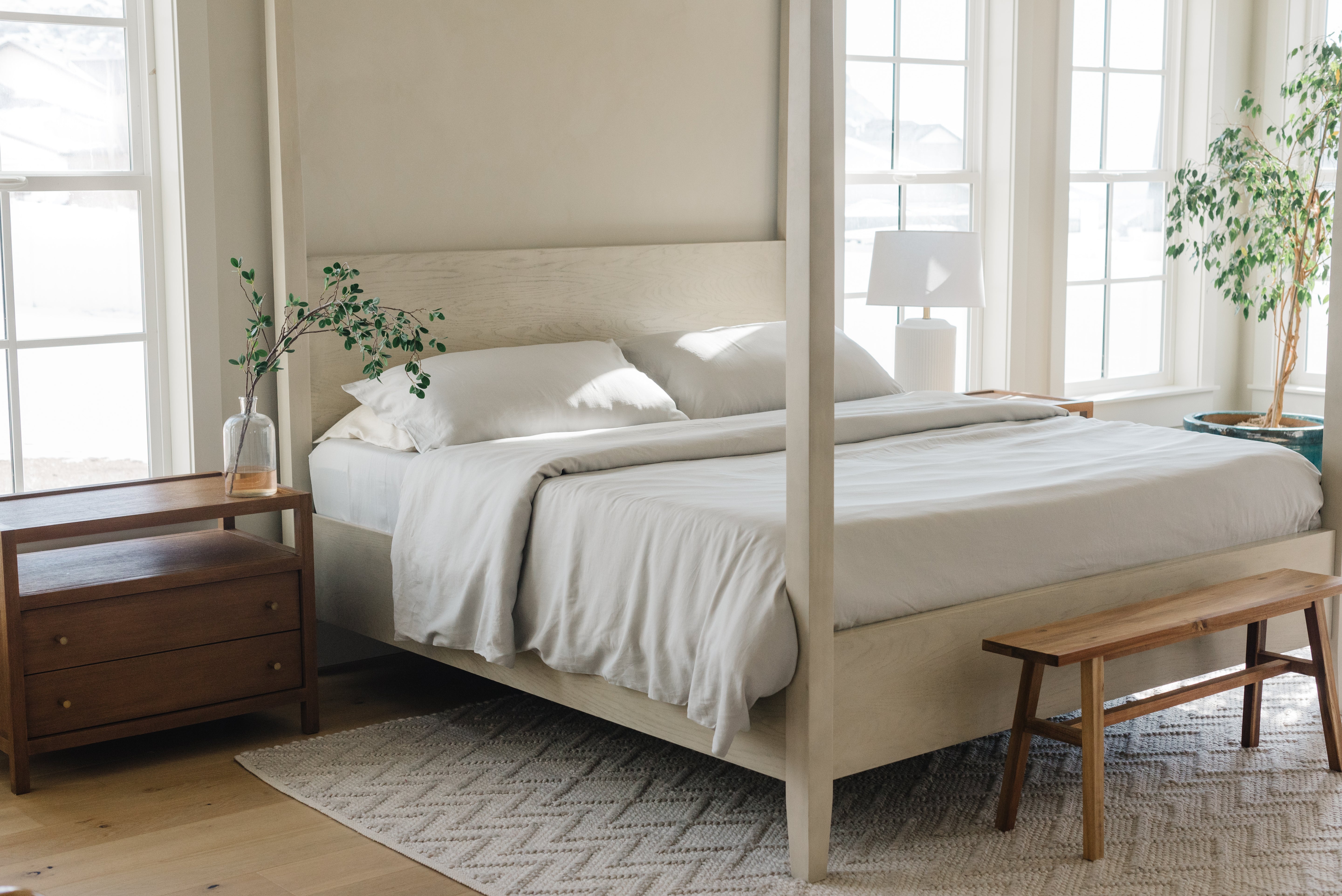 Bright bedroom featuring a white canopy bed with minimalist white bedding, flanked by wooden nightstands and a bench at the foot. Two leafy plants sit by large windows, and natural light streams into the room.