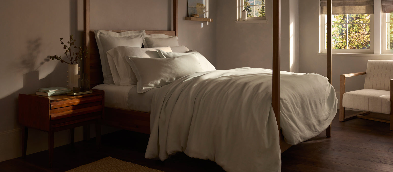 Sunlight streams into a cozy bedroom with a neatly made four-poster bed, white bedding, a wooden nightstand with books and a vase, and a cushioned armchair by the window.