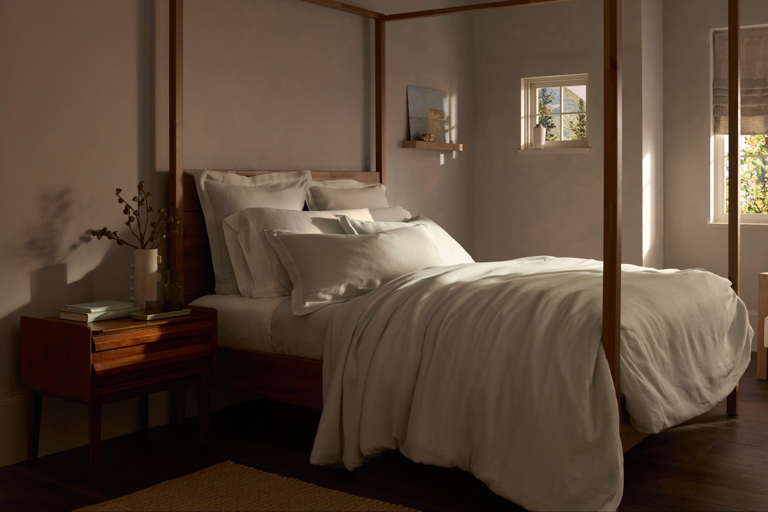 A cozy, sunlit bedroom with a four-poster bed, white bedding, a wooden nightstand with books and a vase, and sunlight streaming through two small windows.