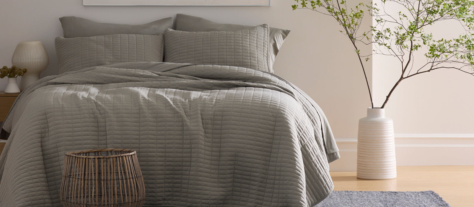 A neatly made bed with gray quilted bedding and pillows in a cozy bedroom, featuring a woven stool, a large vase with branches, and neutral decor.
