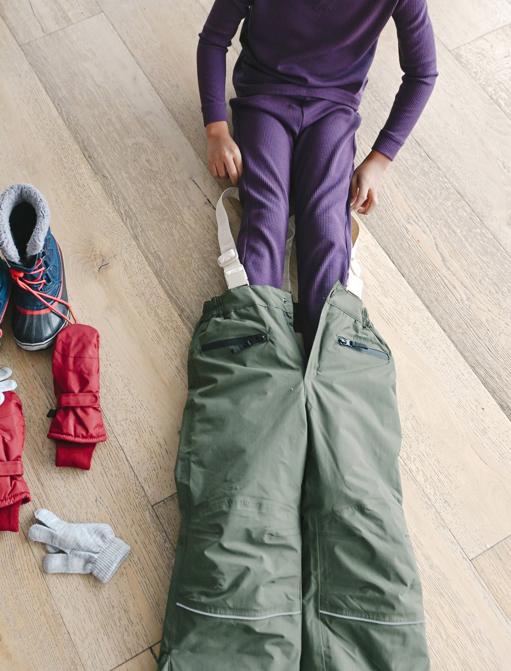 A child wearing Cozy Earth Children's Bamboo Waffle Knit Base Layer Pant in purple sits on a wooden floor, pulling on green snow pants. Nearby are red mittens, gray gloves, and winter boots.