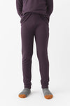 A child stands against a white background wearing Cozy Earth’s Children’s Bamboo Waffle Knit Base Layer Pant in Plum, paired with a matching top and gray socks with orange toes. Their hands are in their pockets, upper body partially out of frame.|Color:Plum