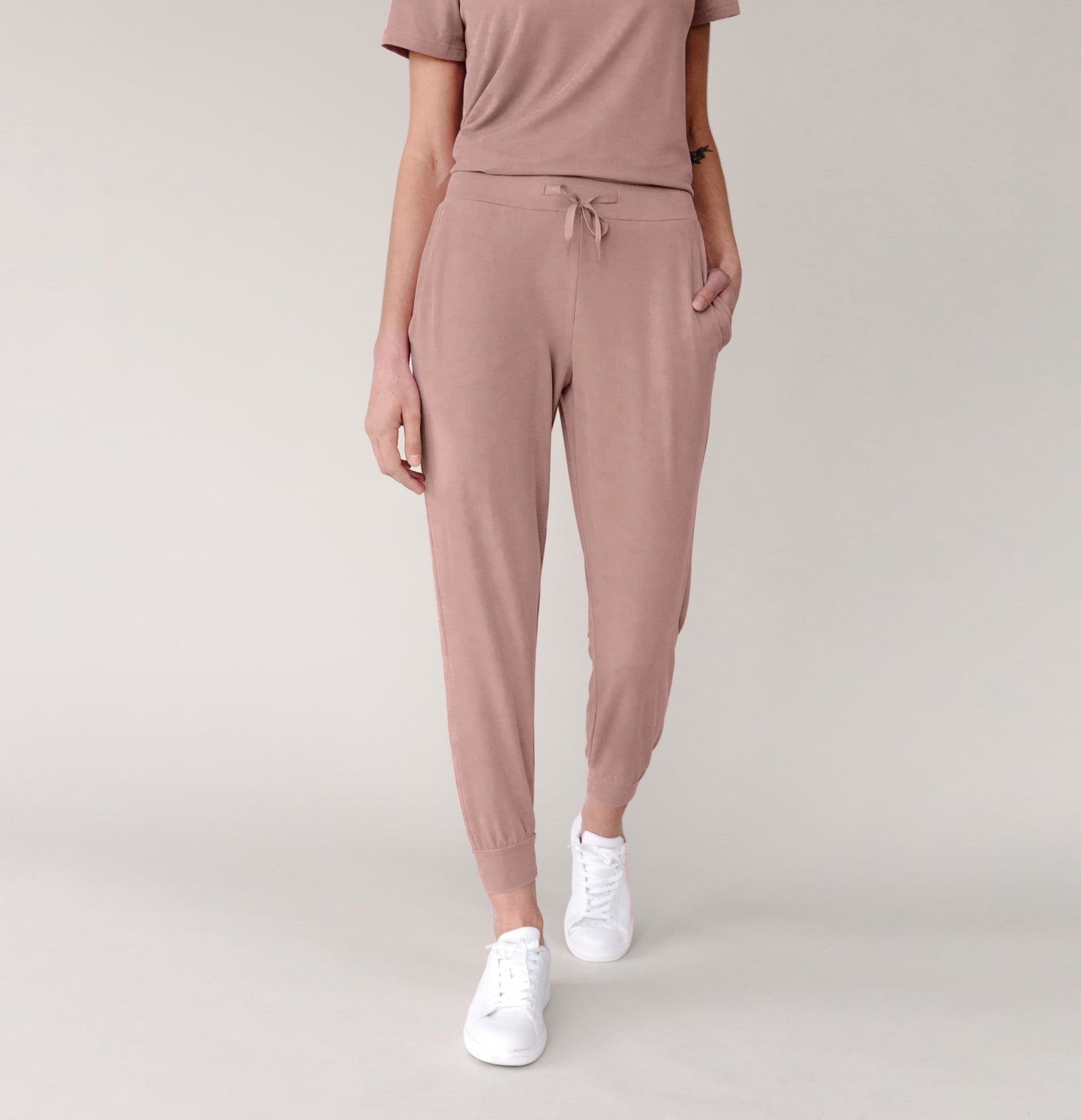 A person is wearing Cozy Earth's Women's Modern Modal Jogger set in Antique Rose. Their right hand is in the jogger pocket, and they are paired with white sneakers. The background is a plain light gray, and the person's head is not visible in the image.