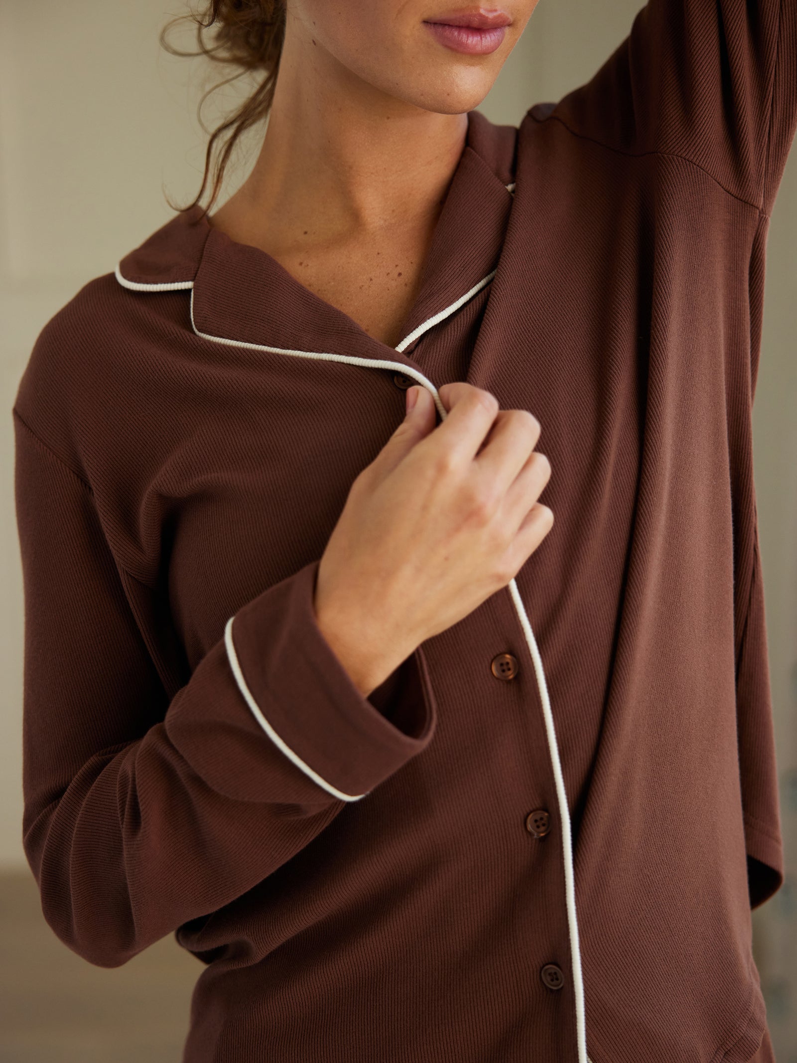 A woman wearing the Cozy Earth Women's Bamboo Rib Knit Classic Long Sleeve Pajama Top in brown with white piping holds her collar while raising her other arm. The image highlights the chest and arm area, with the person's face partially visible. The background is blurred, drawing attention to her attire.