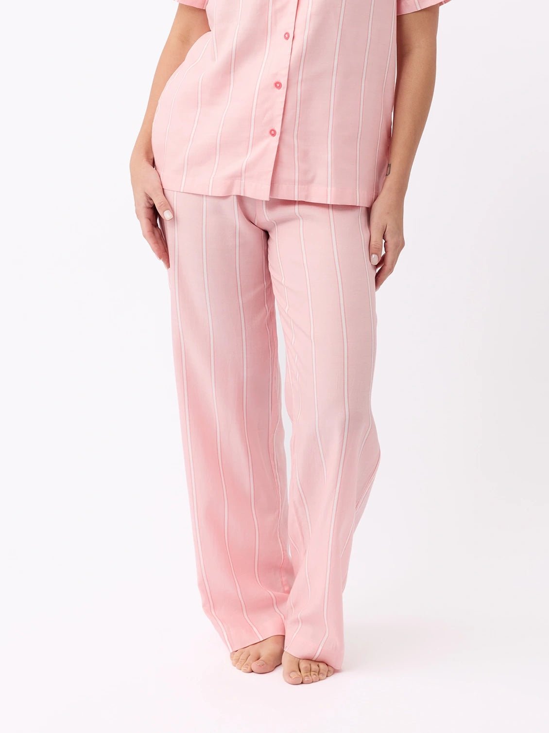 A person stands barefoot on a white background wearing Cozy Earth's Women's Soft Woven Pajama Pant, a light pink, short-sleeved, button-up pajama with white vertical stripes and matching pants.