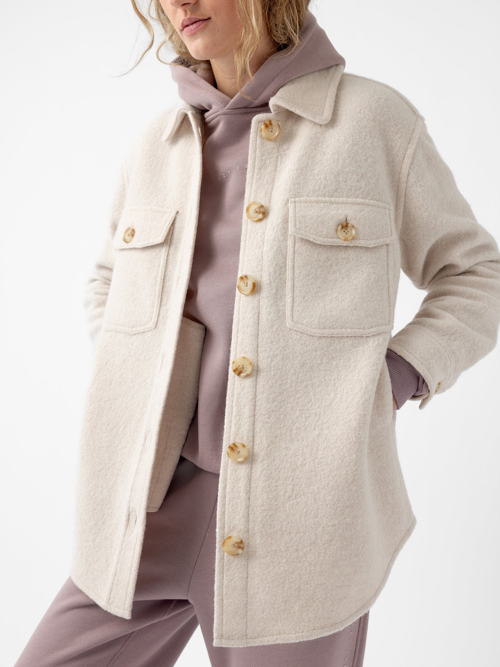 A person wearing a Cozy Earth Women's Boucle Shacket in beige over a mauve hoodie and matching pants. The shacket features large buttons and two front pockets, with their hands tucked into the pockets. The background is plain white.