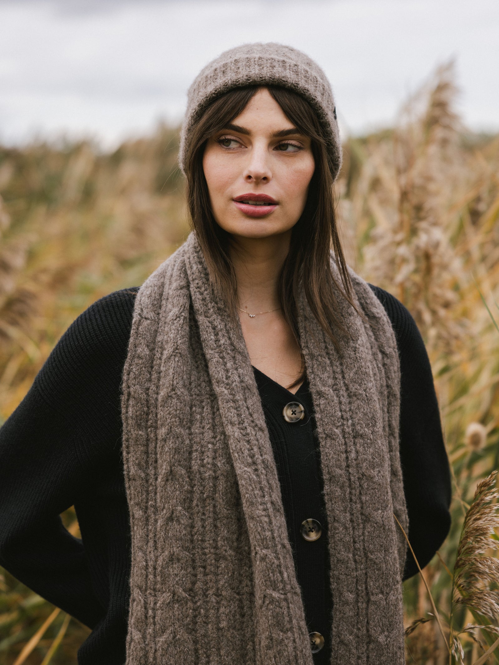 Woman wearing a gray Cozy Earth Cable Knit Beanie and scarf in a field of tall grass.