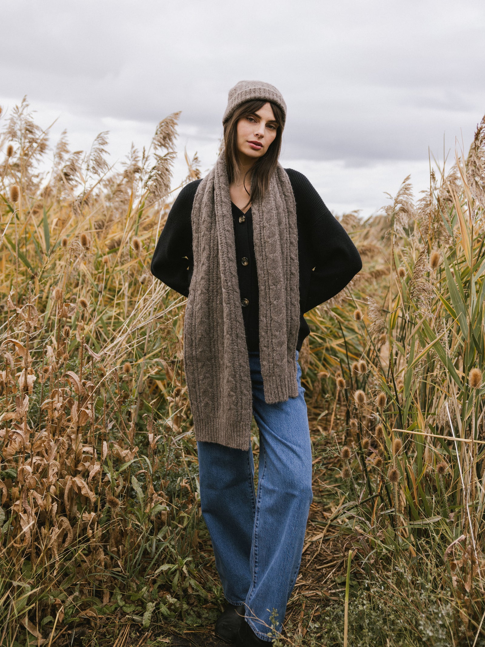 Woman wearing a brown knit cardigan, Cozy Earth Cable Knit Scarf stands in a field of tall grass