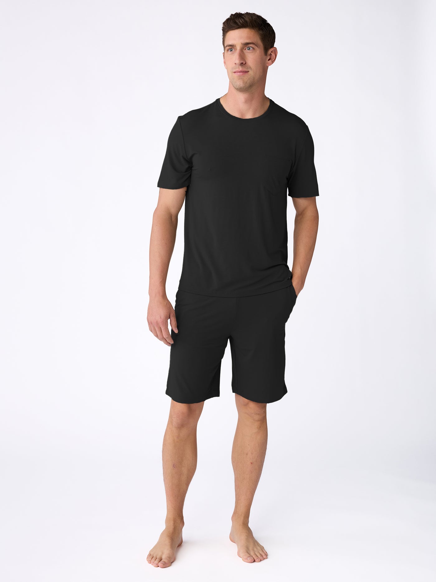 A man stands barefoot against a white background wearing the Cozy Earth Men's Bamboo Stretch-Knit Short Sleeve Pajama Set in black. He has one hand in his pocket and gazes to the side. |Color:Black