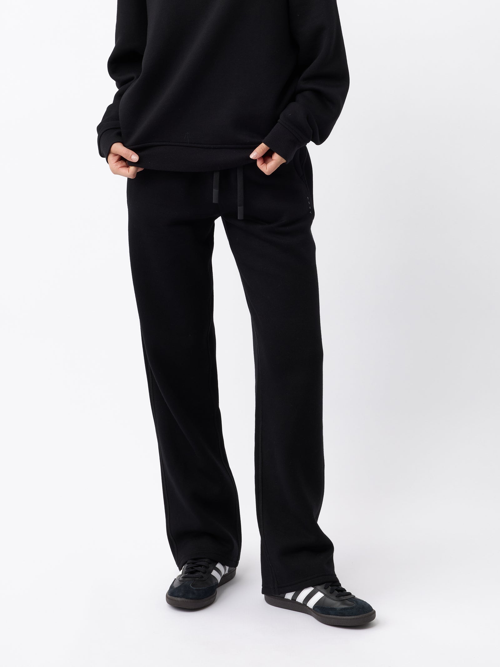 A person is standing against a plain white background, wearing Cozy Earth's Women's CityScape Wide Leg Pant in black and a matching black sweater. They are holding the bottom of their sweater with both hands. Their feet are adorned with black sneakers featuring white stripes.