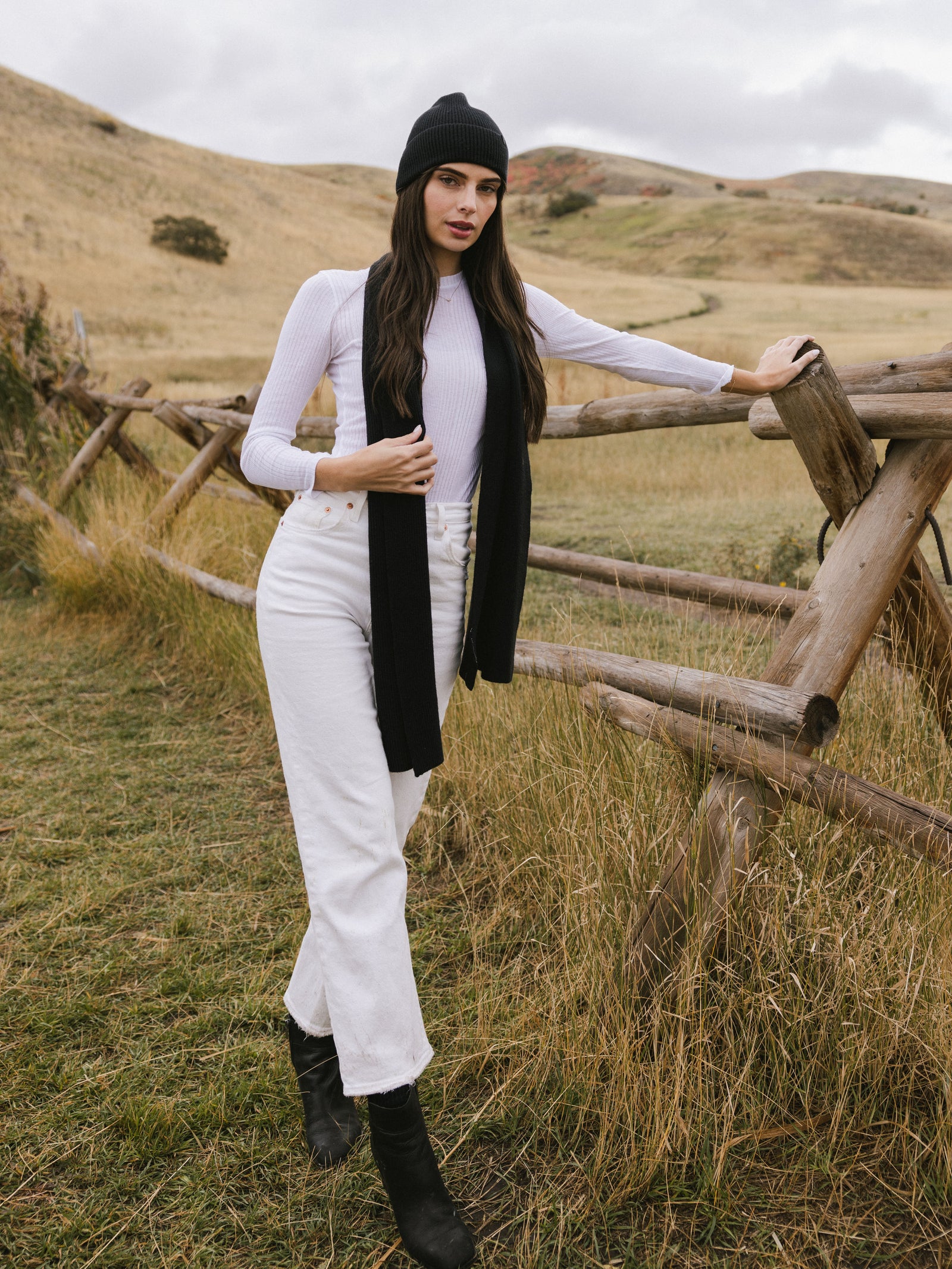 Woman wearing a black scarf, white sweater, and light-colored pants in a field