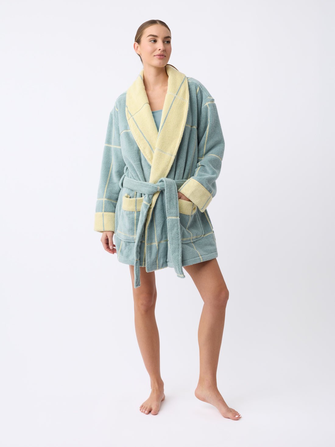 A person wearing a Cozy Earth Windowpane Resort Robe, featuring a blue and yellow checkered pattern with a contrasting yellow collar and a waist belt, stands barefoot against a plain white background. Their hair is tied back.