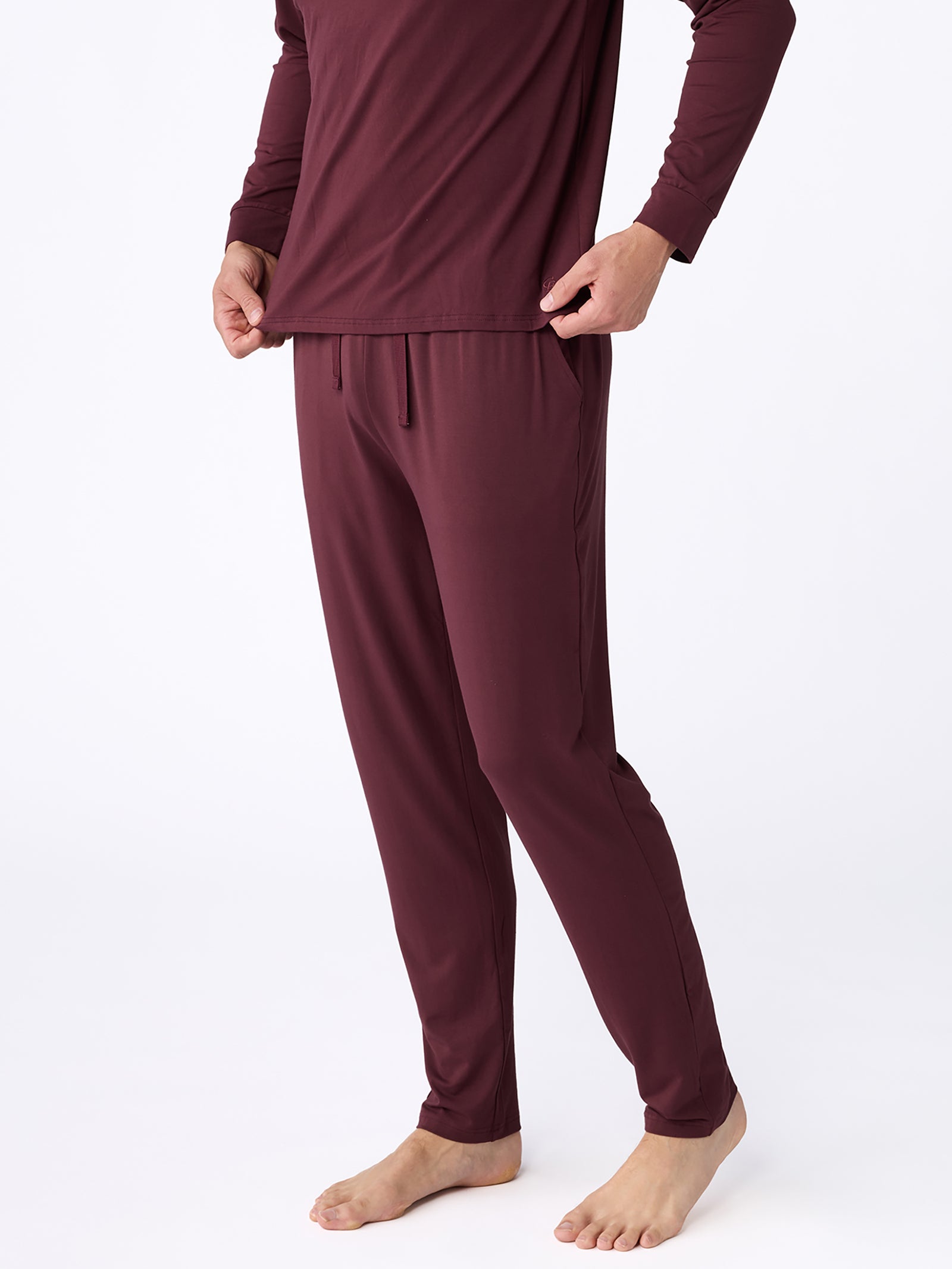 A person wearing the Cozy Earth Men's Stretch-Knit Bamboo Pajama Pant in maroon, along with a matching long-sleeve top, stands barefoot on a white background. The image captures the outfit from the neck down, emphasizing its relaxed fit and soft fabric.