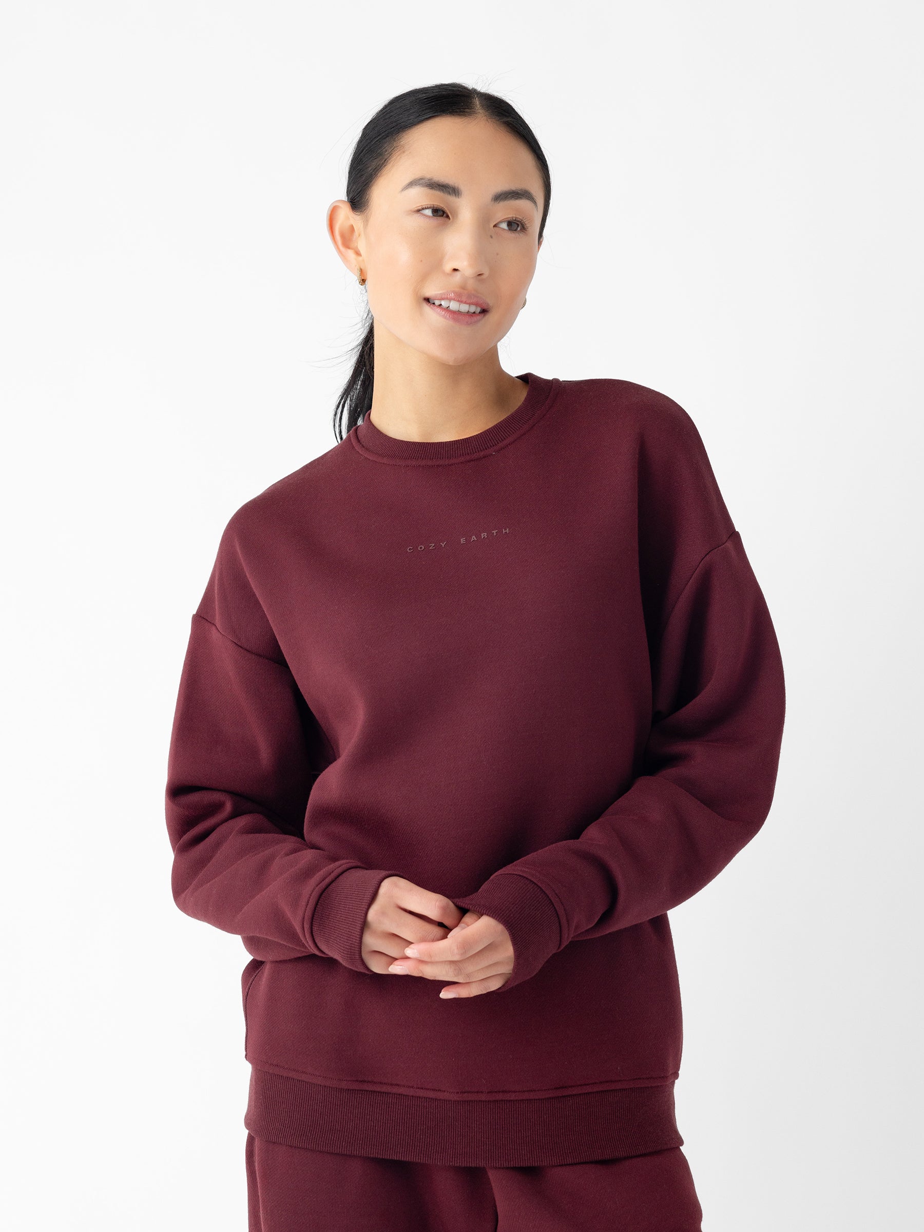 A person with dark hair tied back is wearing the Cozy Earth Women's CityScape Crewneck, paired with matching pants. They are standing against a plain white background. The crewneck features subtle lettering on the front. The person is smiling gently and has their hands clasped together. |Color:Burgundy