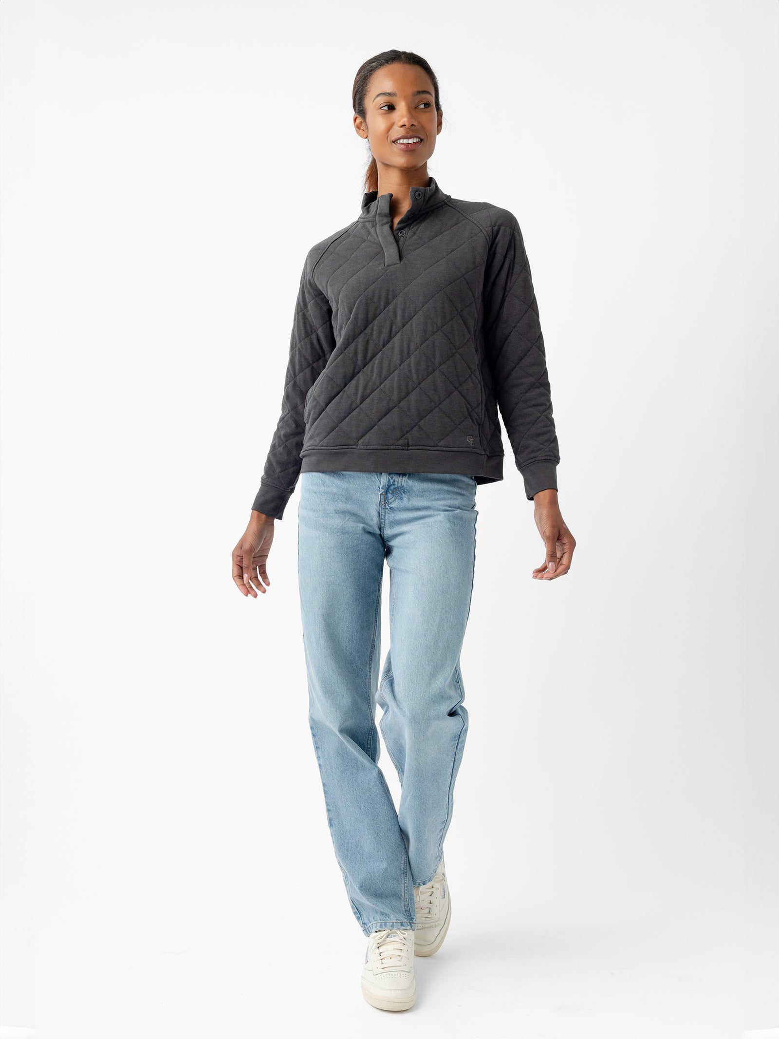A person with long dark hair is walking while looking slightly to the right. They are wearing a Women's Ultra-Soft Bamboo Quilted Snap Pullover by Cozy Earth, light blue jeans, and white sneakers. The background is a plain white studio setting.