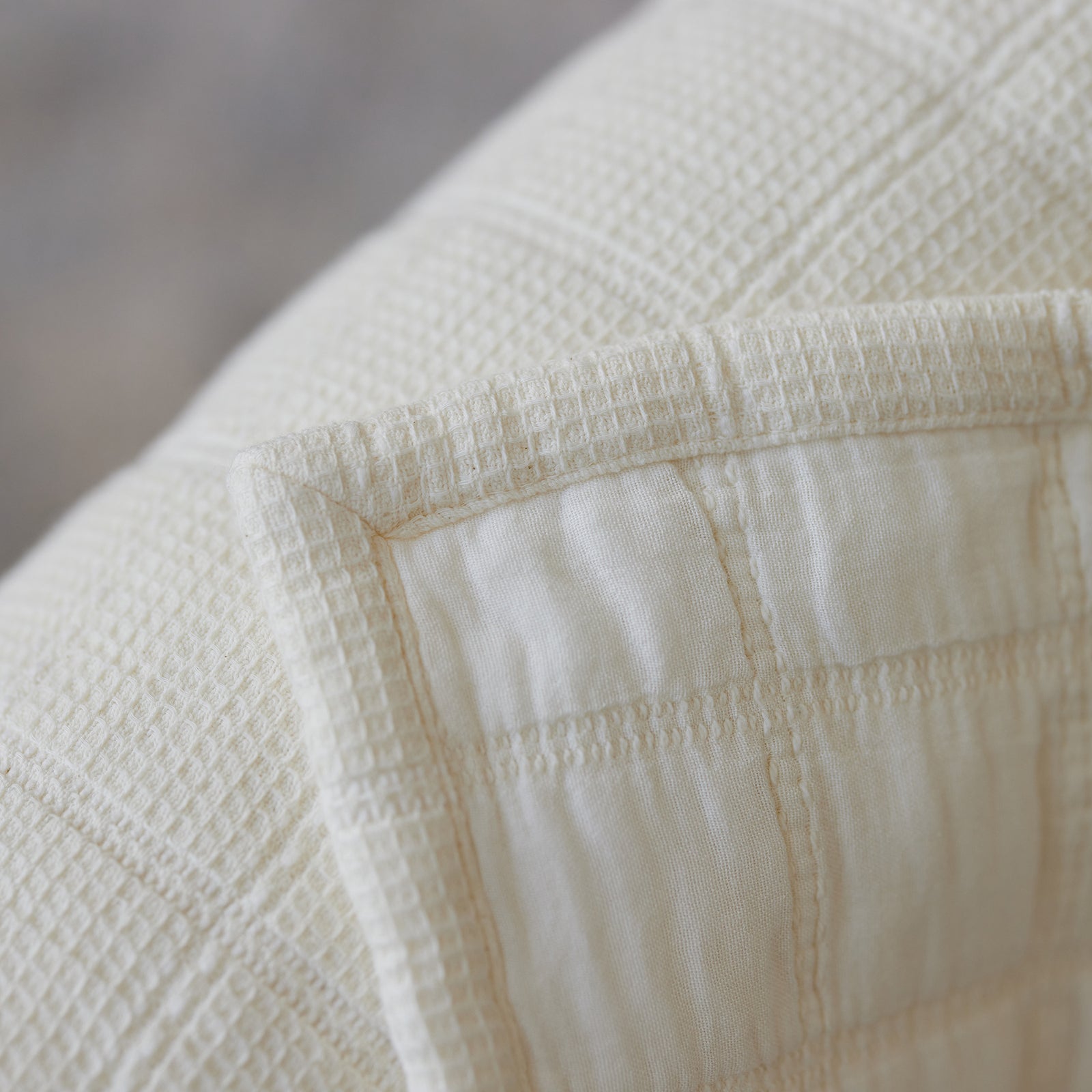 Close-up of the Waffle Windowpane Blanket by Cozy Earth. This cream-colored blanket showcases a textured fabric with a checkerboard pattern and subtle grid design. The meticulous stitching detail along the edges gives it a neat and tailored appearance.