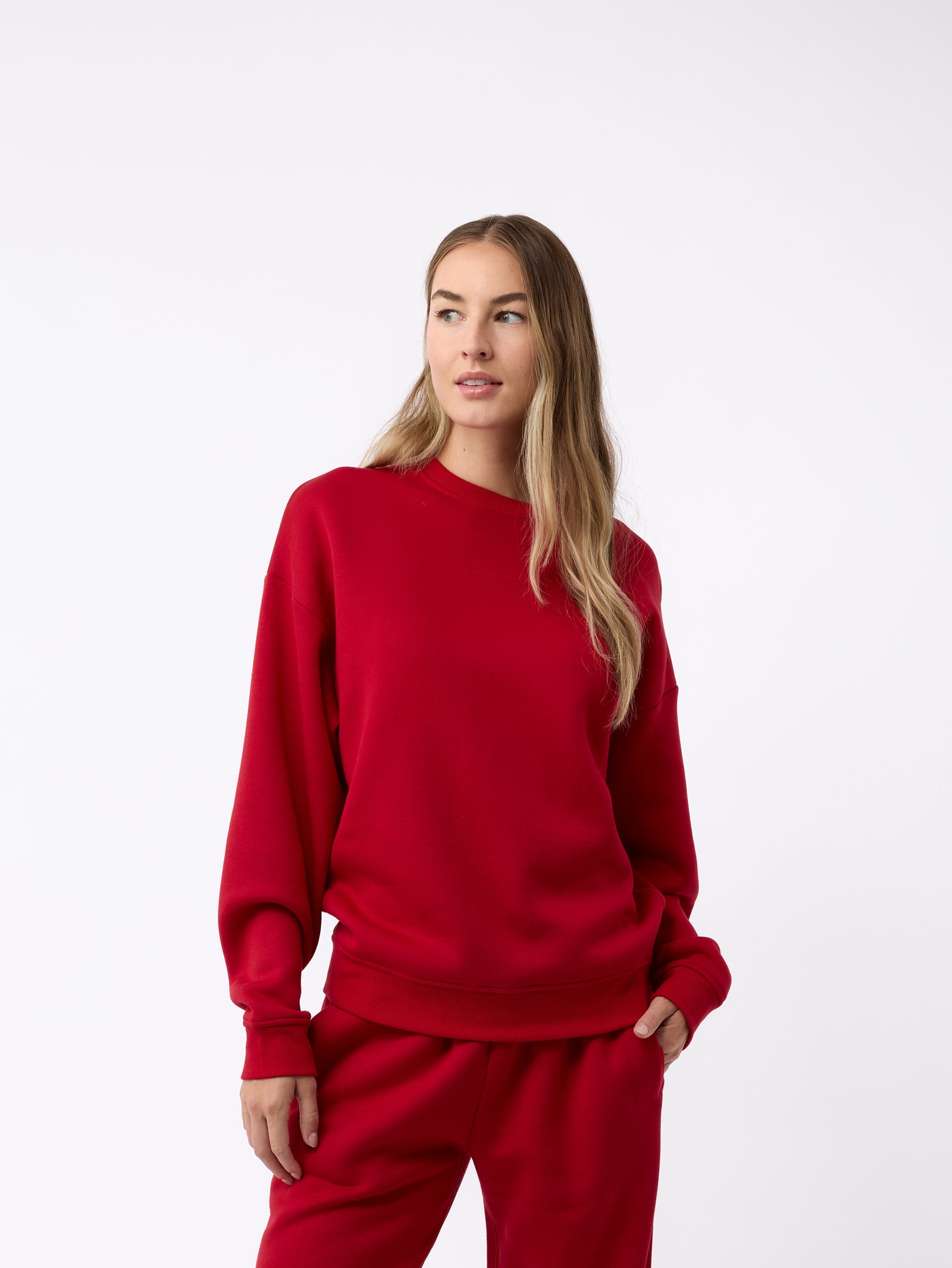 A person with long hair is wearing a Cozy Earth Women's CityScape Crewneck and matching sweatpants, standing against a plain white background. They have one hand in their pocket and are gazing slightly to the side.