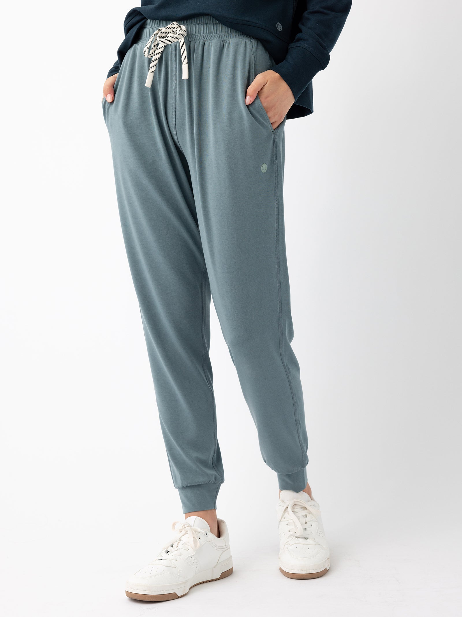 A person is standing against a plain white background, modeling the Women's Studio Jogger from Cozy Earth, which are loose-fitting, light blue jogger pants featuring an elastic waistband and drawstring. They are also wearing a dark long-sleeve top and white sneakers with a thick sole. Hands are in the pockets.