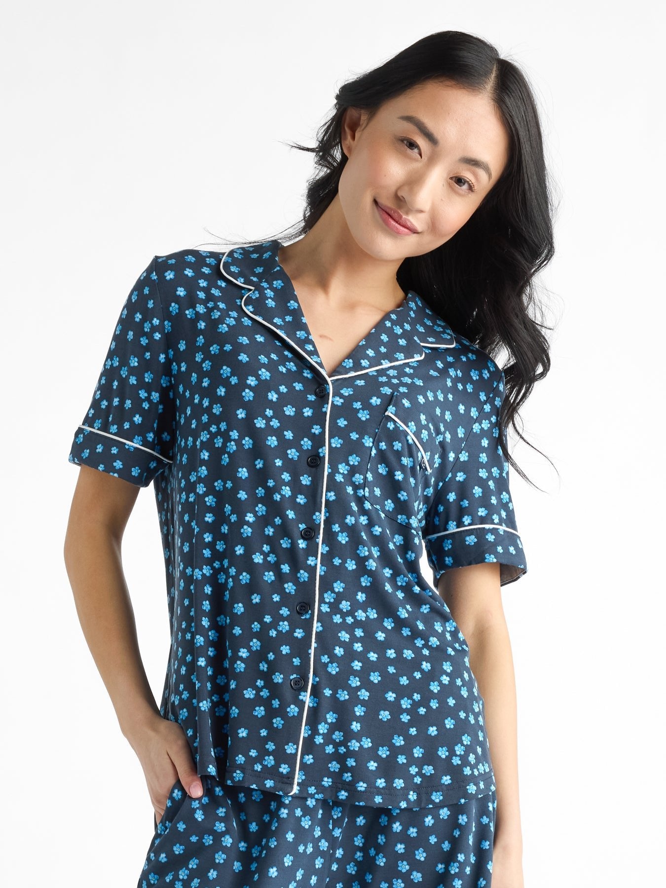 A person wears the Women's Bamboo Stretch-Knit Short Sleeve Pajama Top by HIDE, featuring dark blue with light blue floral patterns. They stand against a white background, smiling with one hand in a pocket. The top has a collar and buttons.