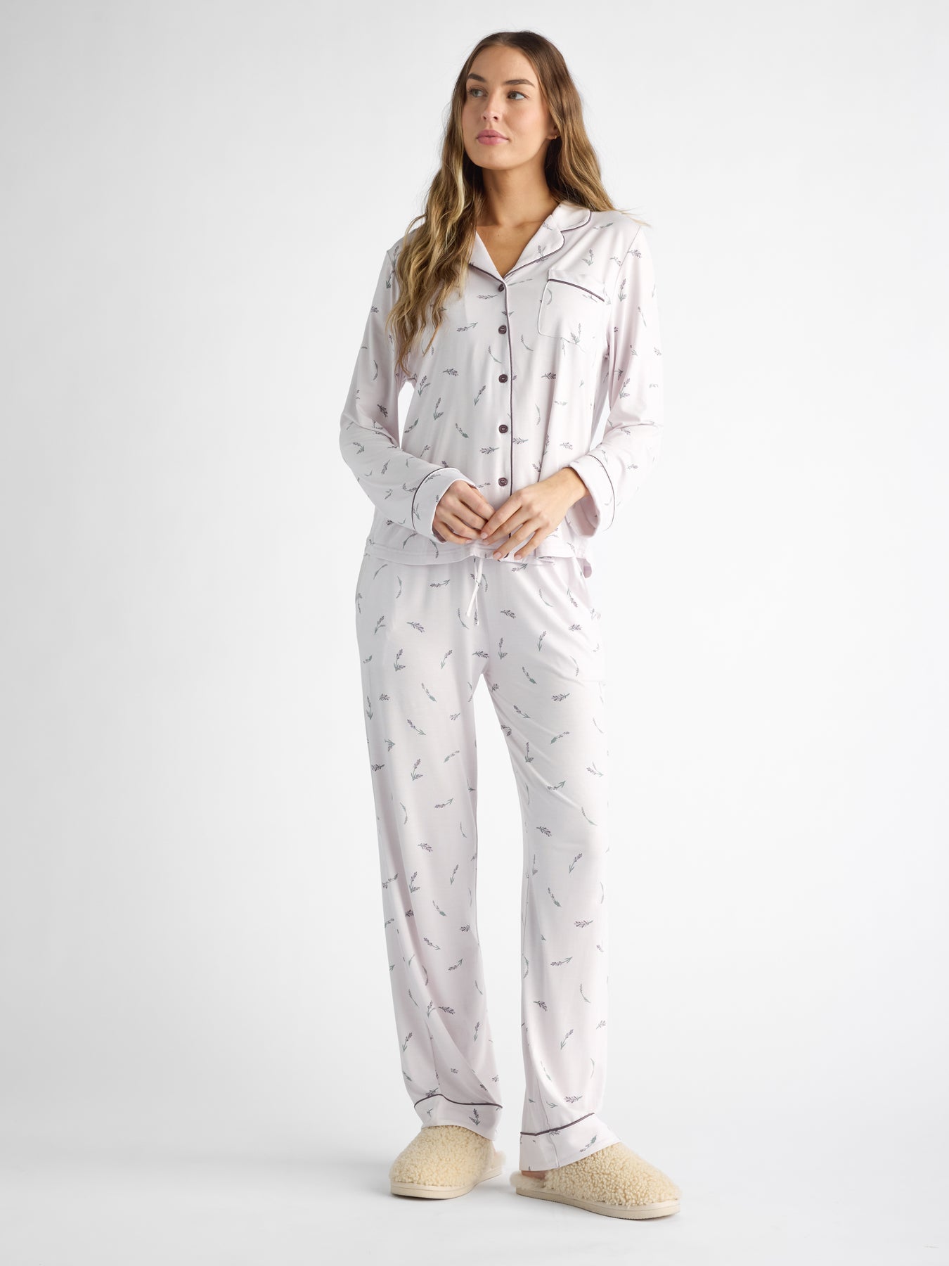 A person wearing the Women's Bamboo Stretch-Knit Long Sleeve Pajama Top by Cozy Earth, styled with buttons and a collar, stands in subtle-patterned white pajamas against a plain backdrop. They sport pajama pants with a drawstring and beige slippers.