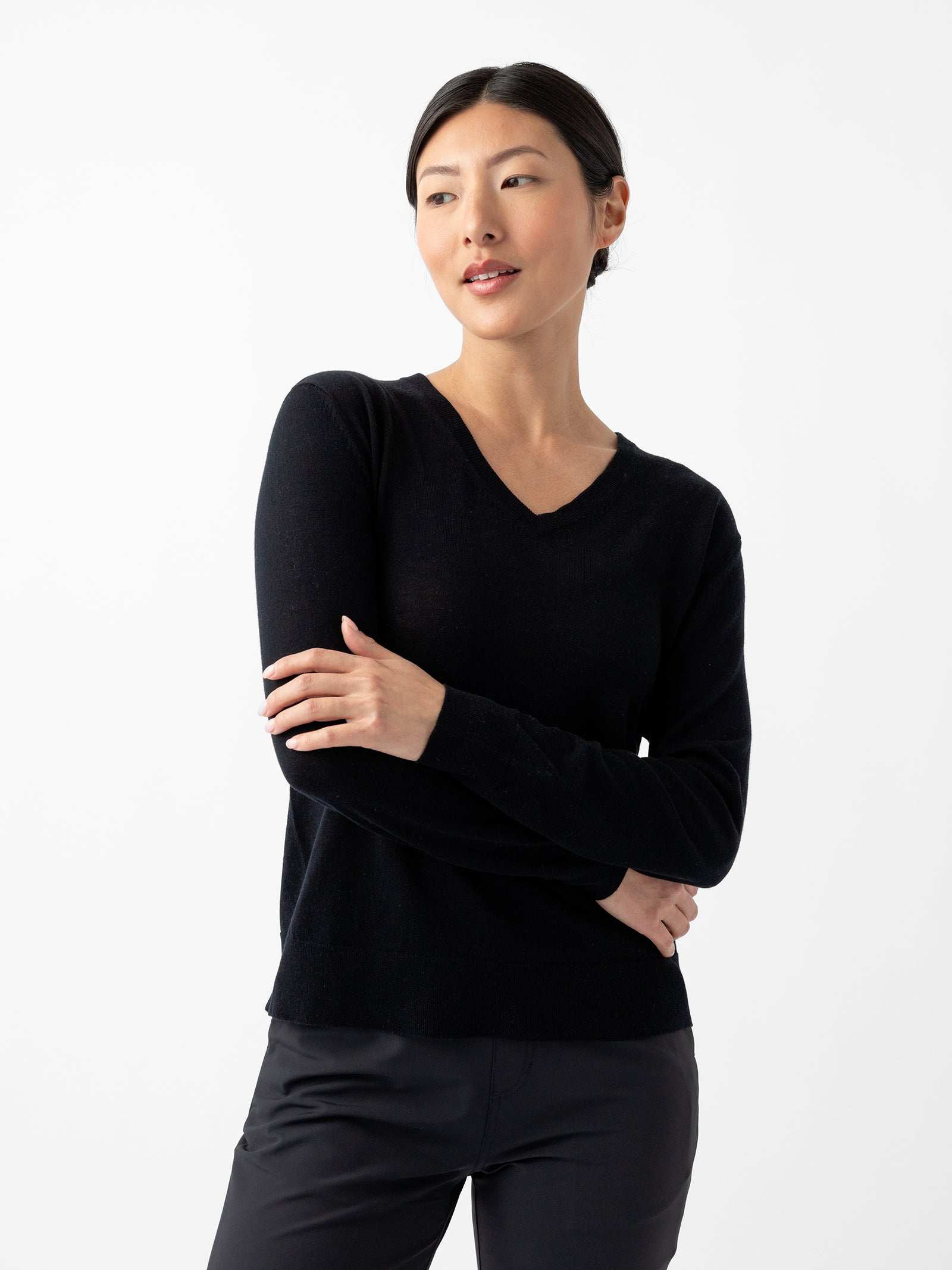 A woman with dark hair is wearing a Cozy Earth Women's AirKnit V-Neck Sweater in black along with black pants. She is standing with her arms crossed and looking slightly to the side with a neutral expression. The background is plain white.