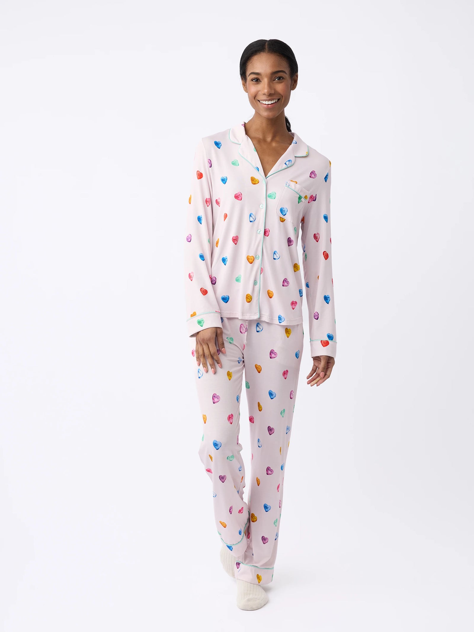 A person smiles in a Cozy Earth Women's Stretch-Knit Bamboo Pajama Set, featuring colorful heart patterns. The shirt has long sleeves with a collar, paired with loose-fitting pants against a plain white background.