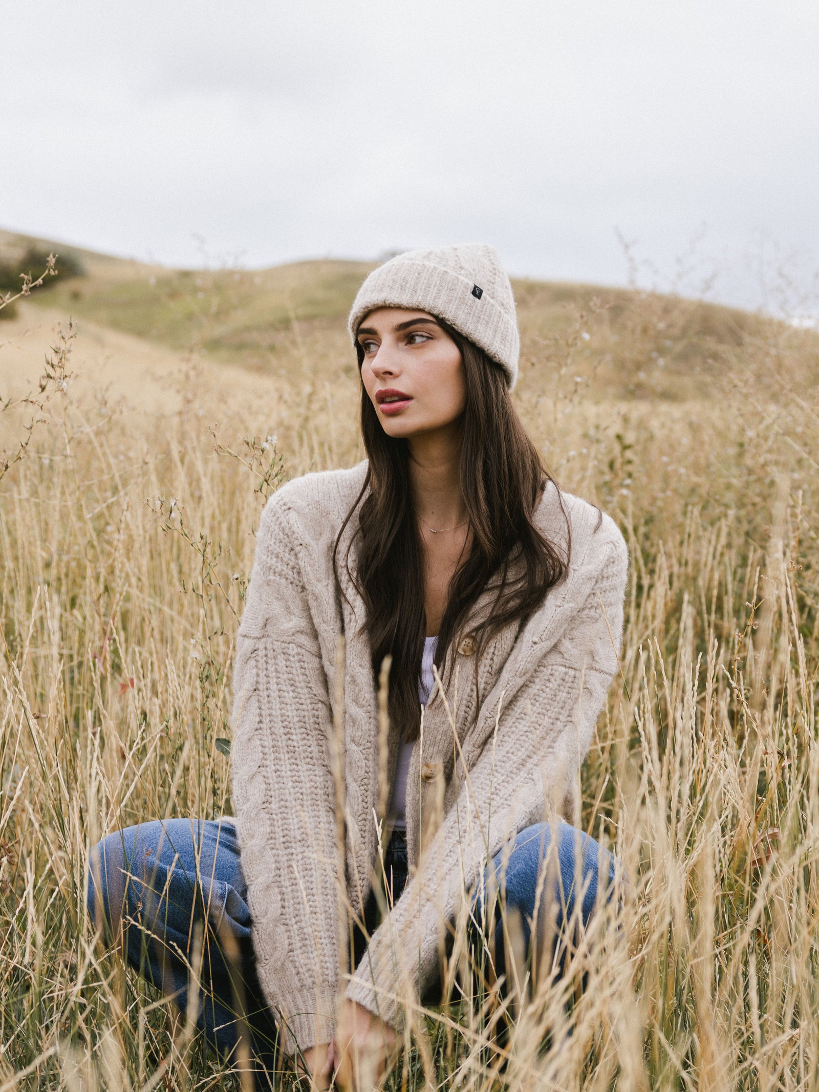 Woman wearing a tan Cozy Earth Cable Knit Beanie and scarf in a field of tall grass.