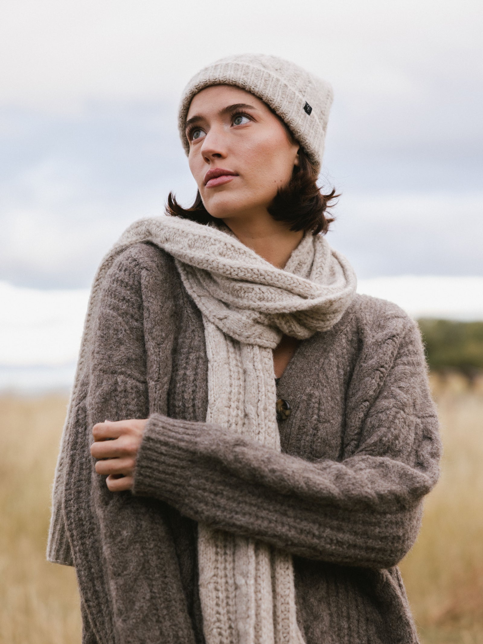 Woman wearing a brown knit cardigan, Cozy Earth Cable Knit Scarf in beige, and a beanie in a field.