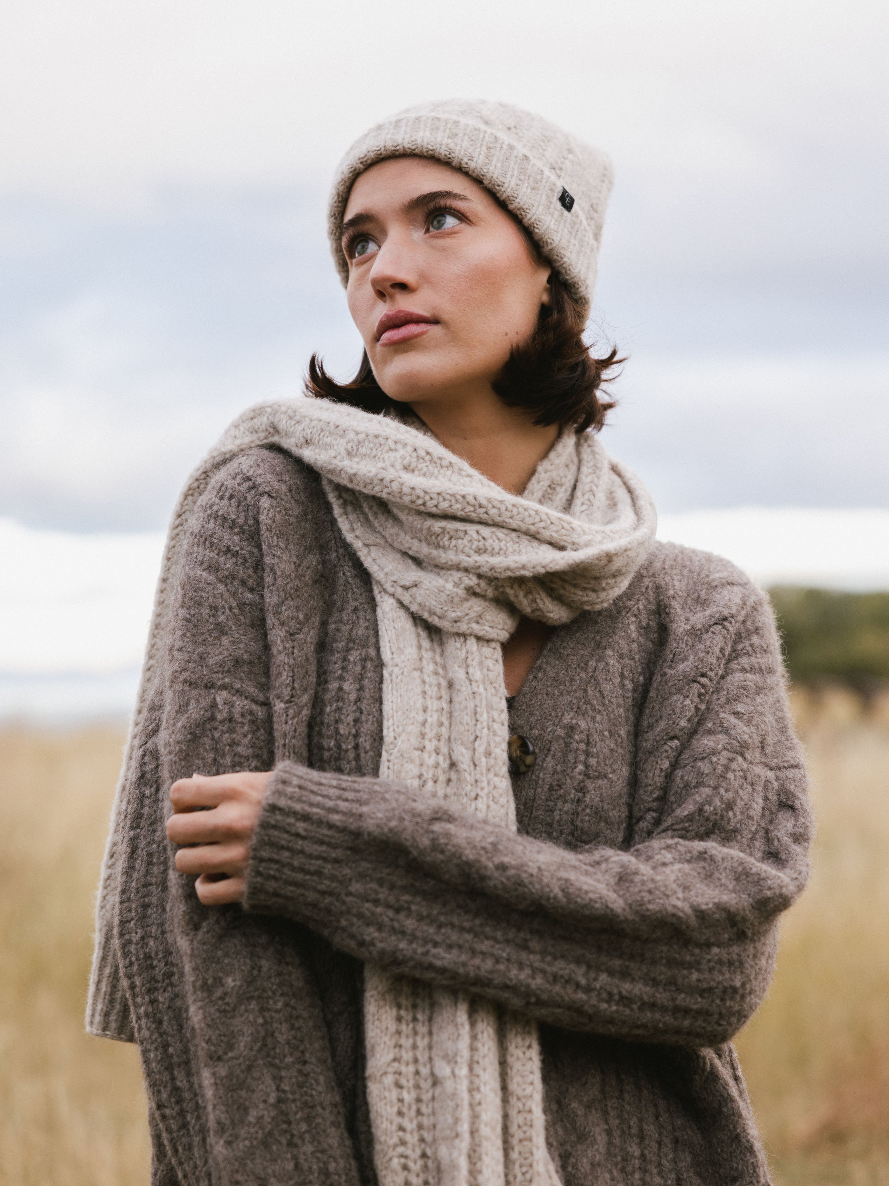 Woman wearing a brown knit cardigan, Cozy Earth Cable Knit Scarf in beige, and a beanie in a field. |Color:Oatmeal