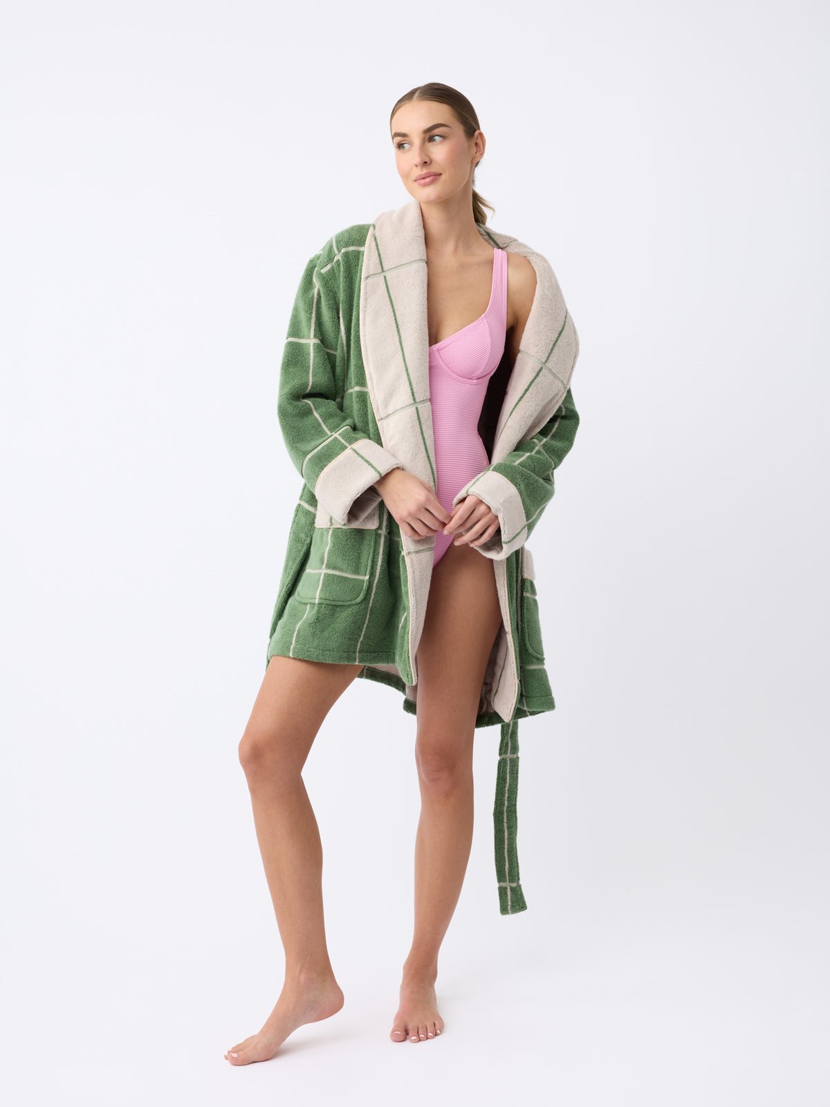 A person stands barefoot against a plain white background, wearing Cozy Earth's Windowpane Resort Robe in green and white checkers over a pink swimsuit. They have long hair and maintain a relaxed sideward gaze.