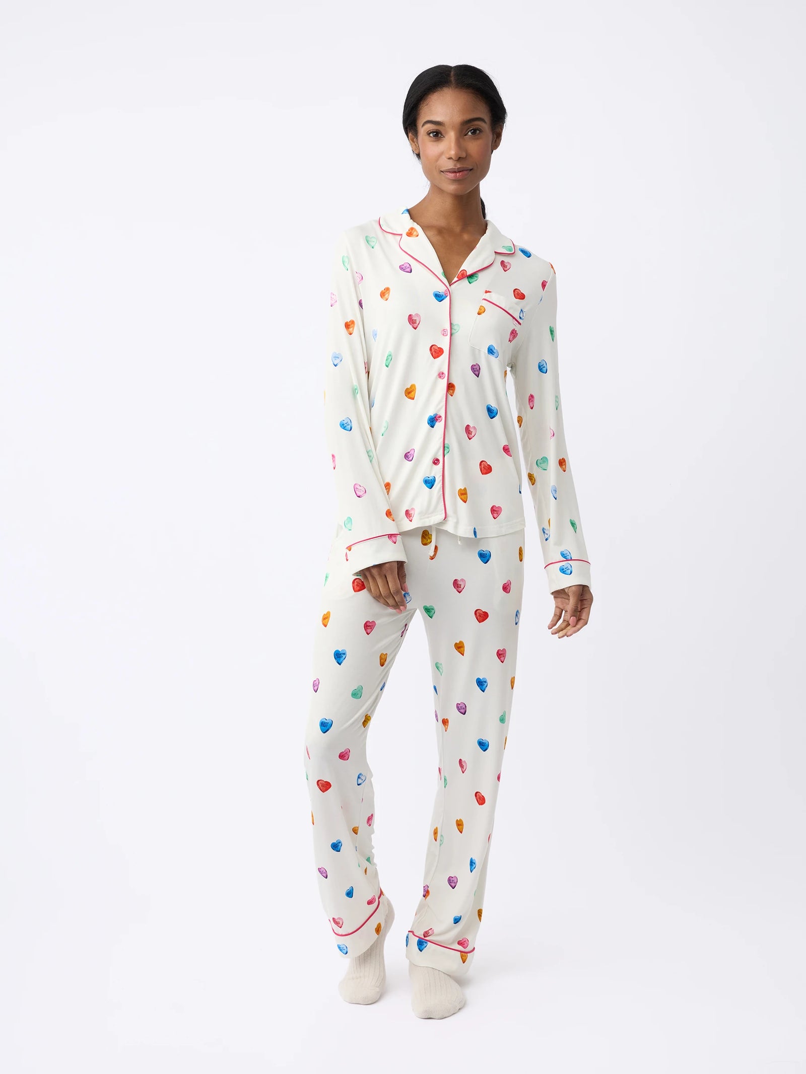 A person wears a white Women's Stretch-Knit Bamboo Pajama Pant by Cozy Earth with colorful heart patterns, featuring a button-up shirt with a collar and matching pants, against a plain background. They also wear light-colored socks.
