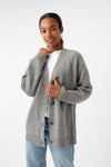 A person is styled in an Oversized Sunday Cashmere Cardigan from Cozy Earth, layered over a white shirt and blue jeans. Standing against a plain white background, they are slightly smiling with one hand gently adjusting the cardigan. |Color:Speckled Grey