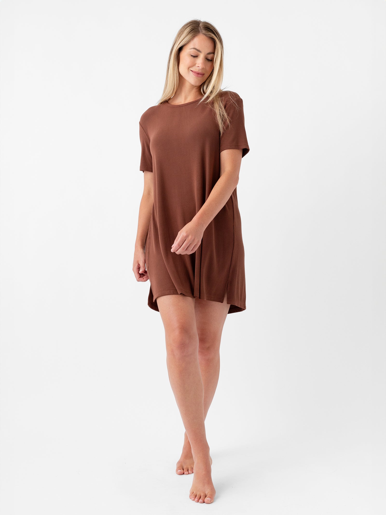A woman with long blonde hair stands barefoot against a white background, wearing the Cozy Earth Women's Bamboo Rib-Knit Boyfriend Sleep Dress. She looks down, holding the hem of the loose-fitting, short-sleeved brown dress gently with one hand.