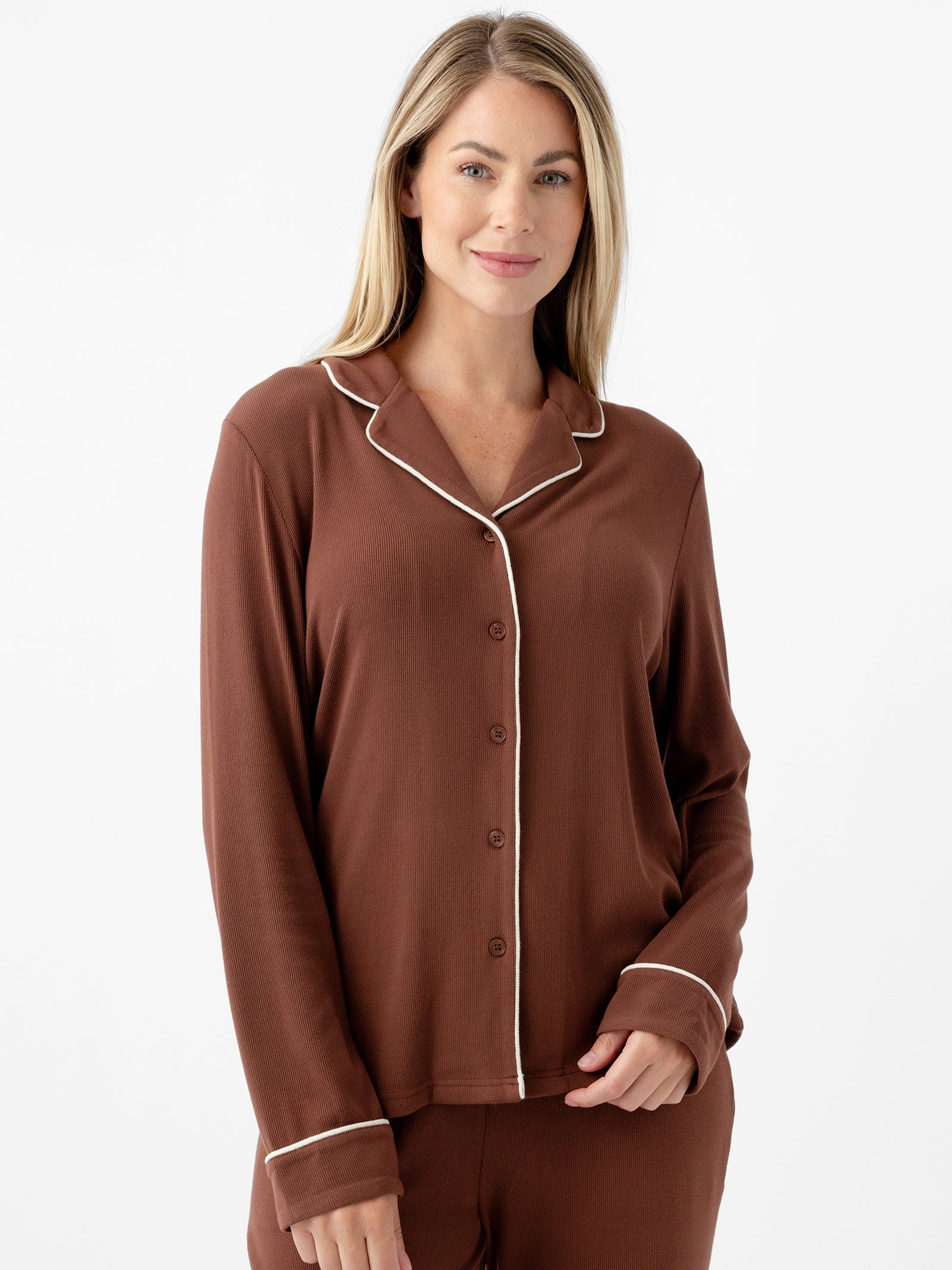 A person with long blonde hair stands against a plain white background, wearing a brown Women's Bamboo Rib Knit Classic Long Sleeve Pajama Top by Cozy Earth. The pajama top features a buttoned front, a collar, and white piping along the edges. The person is smiling softly and has one hand in their pocket.