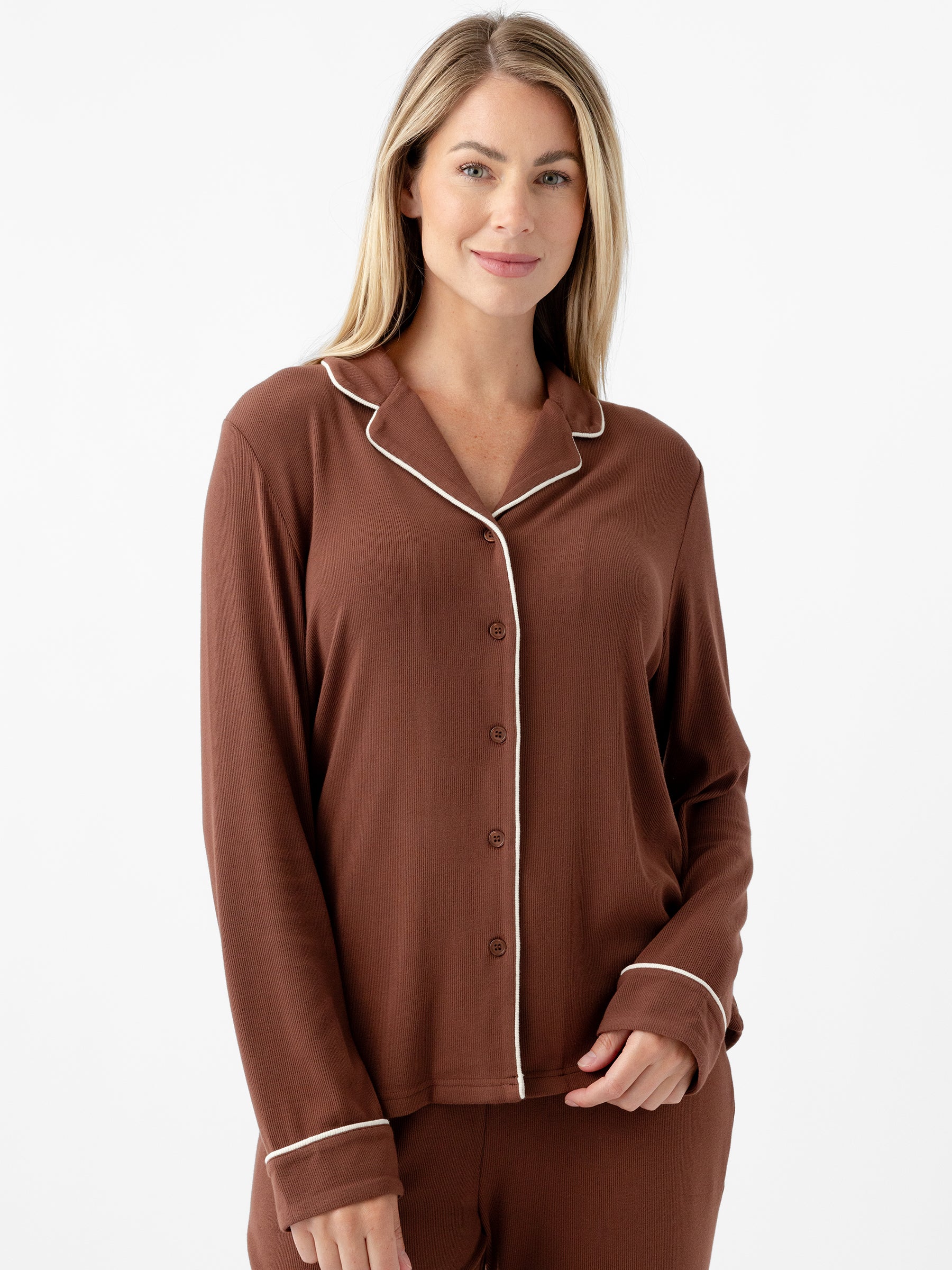 A person with long blonde hair stands against a plain white background, wearing a brown Women's Bamboo Rib Knit Classic Long Sleeve Pajama Top by Cozy Earth. The pajama top features a buttoned front, a collar, and white piping along the edges. The person is smiling softly and has one hand in their pocket. |Color:Spice