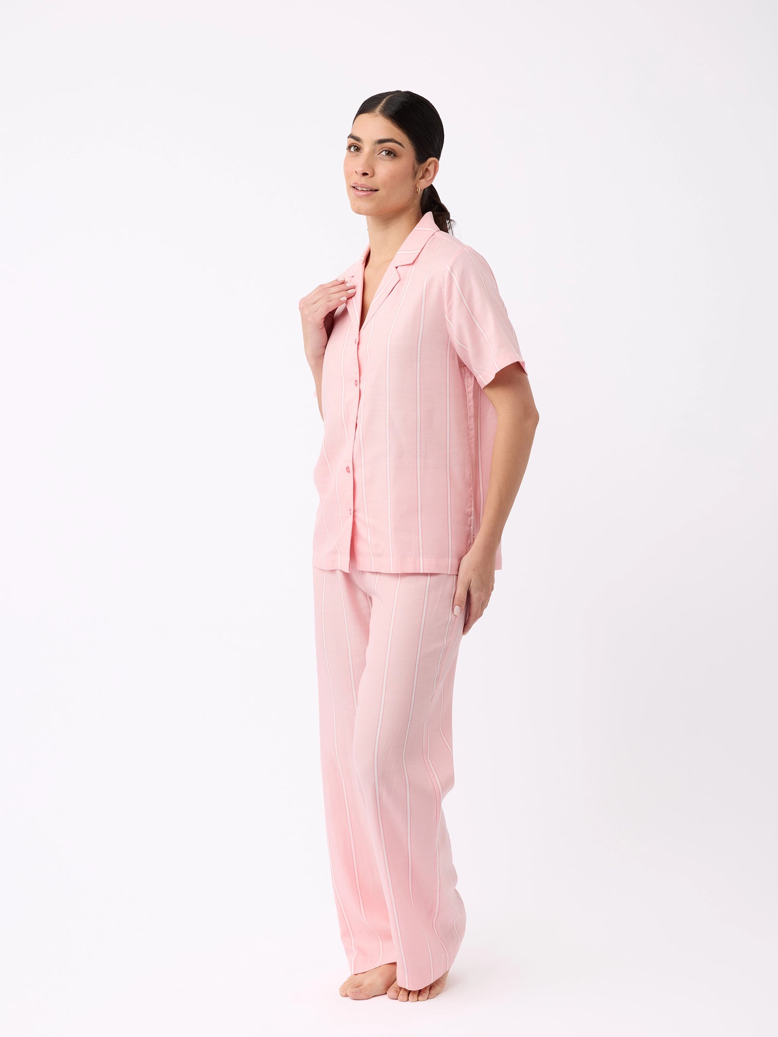 A person with long dark hair tied back smiles gently while standing barefoot against a white background, wearing Cozy Earth's Women's Soft Woven Pajama Pant set in pink with white pinstripes, featuring a matching short-sleeve button-up shirt and long pants.