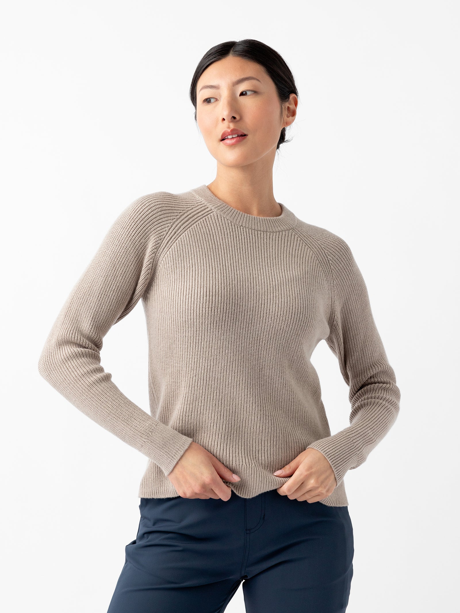 A person stands against a plain white background, wearing Cozy Earth's Women's Classic Crewneck in beige and dark blue pants. The individual has dark hair tied back and is looking to the side with hands resting on the hips.