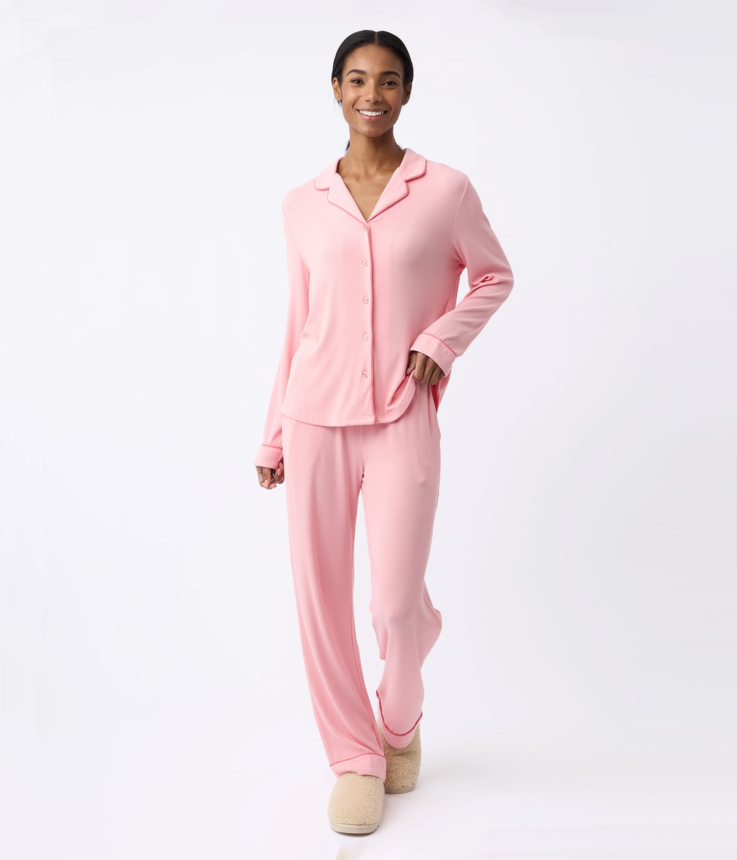 A person wearing the Cozy Earth's Rib-Knit Classic Long Sleeve Pajama Set in light pink with matching pants and a button-up top smiles while standing in light beige slippers against a white background.