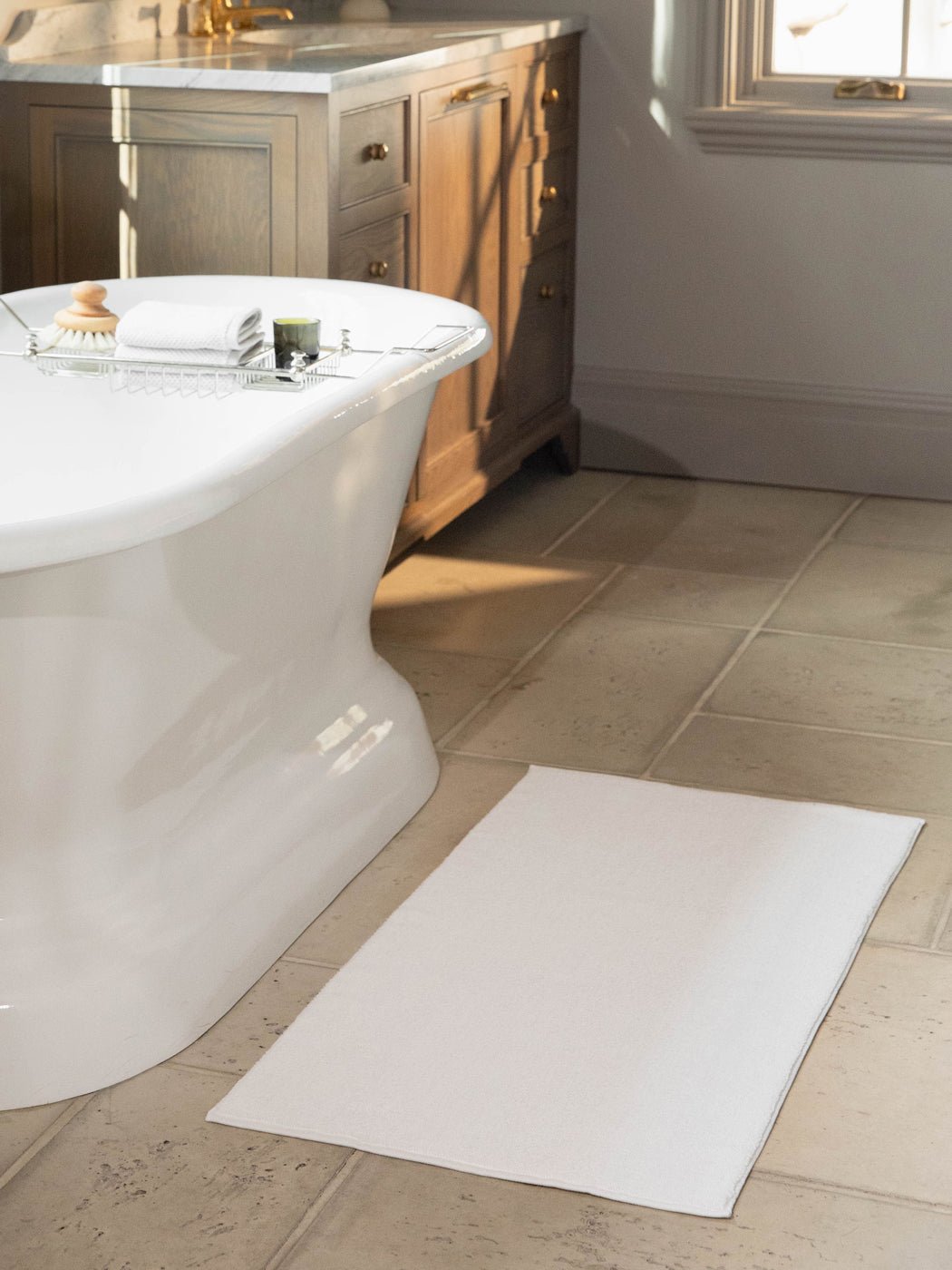 The Cozy Earth Loop Terry Bath Mat in white rests on a beige tiled floor beside a freestanding tub, with a wooden vanity with drawers and a window visible in the background.