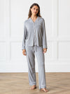 A woman is standing on a wooden floor in a room with white paneled walls, dressed in the Women's Bedtime Bundle by Cozy Earth. The gray pajama set features white trim, with the top boasting buttons and a front pocket, while the pants include a drawstring waist. Her long hair flows freely as she stands barefoot. |Color:Grey
