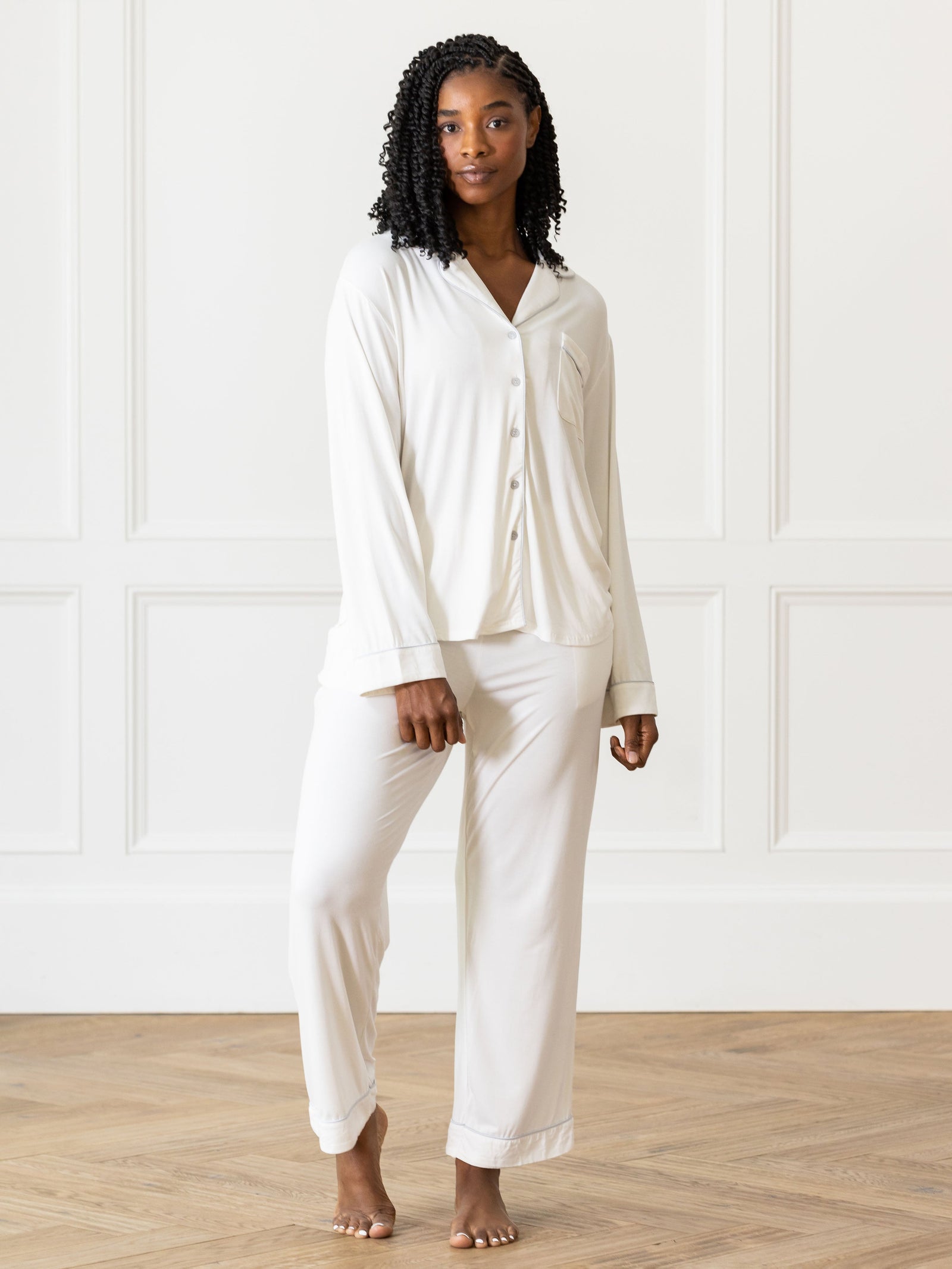 A person with braided hair is standing on a wooden floor against a white paneled wall, wearing the Cozy Earth Women's Bedtime Bundle, which consists of a cream-colored long-sleeved button-up top and matching pants.