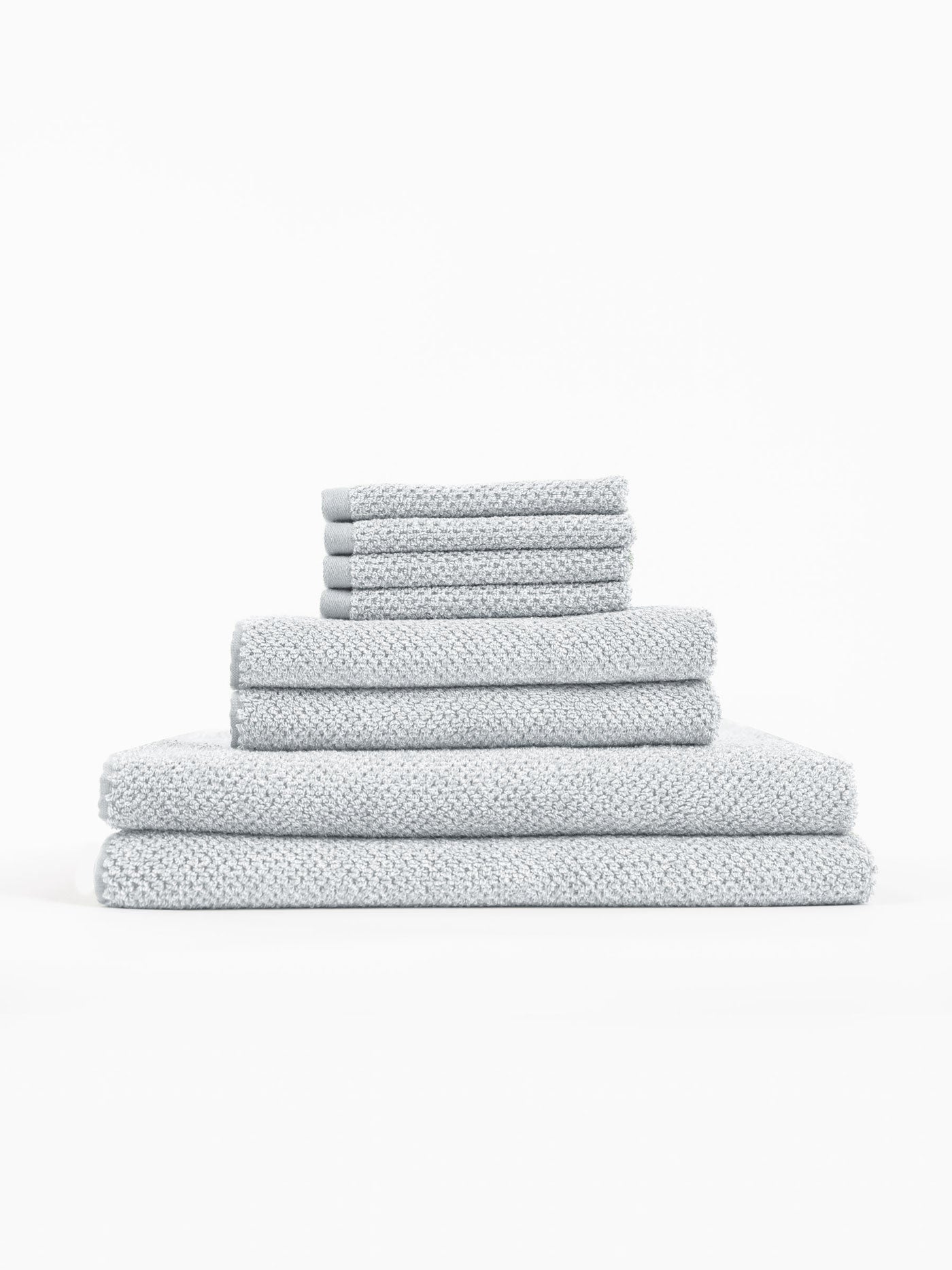 The Cozy Earth Nantucket Bath Towel Set features light grey textured towels in multiple sizes, neatly stacked from largest to smallest against a plain white background.|Color:Heathered Harbor Mist