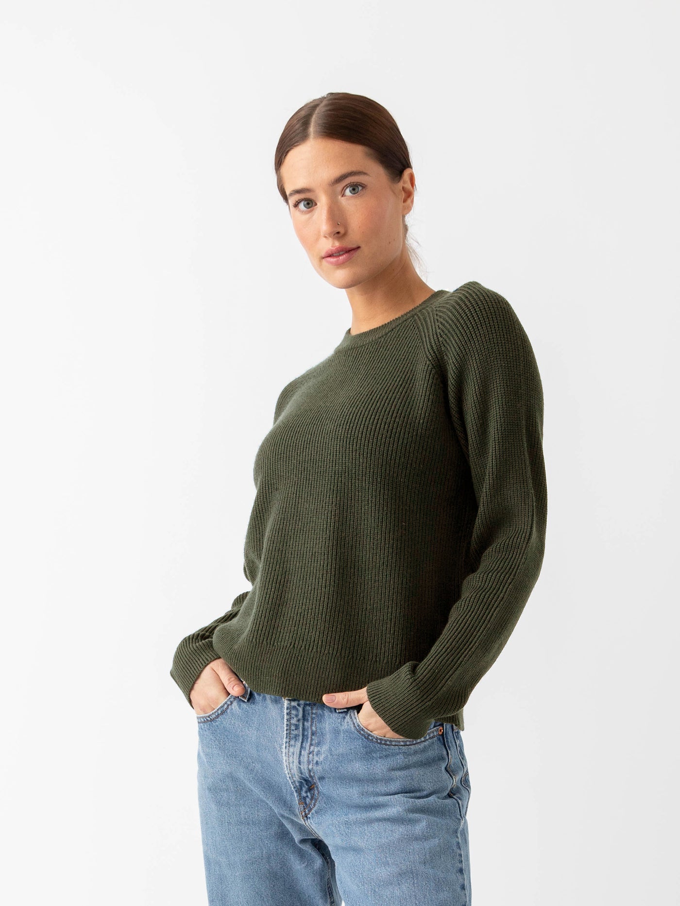 A person with long hair is wearing a Cozy Earth Women's Classic Crewneck in dark green and blue jeans, standing against a plain white background. Their hands are in their pockets, gazing directly at the camera.