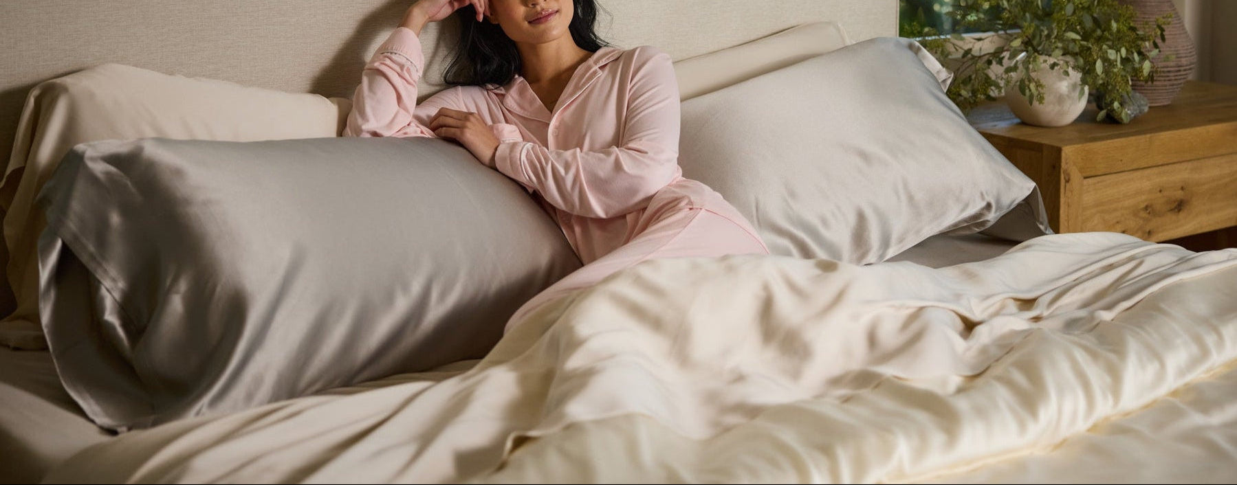 A woman in pink pajamas relaxes on a bed with light-colored bedding, leaning against large pillows and resting her head on her hand. A nightstand with a lamp and a potted plant is visible beside the bed.