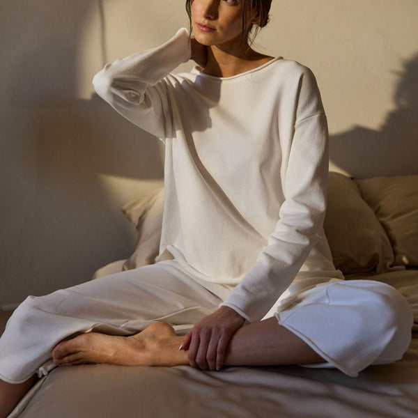 A person sits cross-legged on a bed wearing Cozy Earth's Women's Chelsea Pullover, gazing thoughtfully to the side with one hand on their neck. Warm sunlight casts gentle shadows on the wall behind them. |Color:Bone