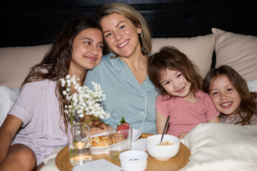 A woman and two children smile while sitting closely together on a sofa. A wooden tray with breakfast items like croissants and yogurt is in front of them. There are small white flowers in a vase on the tray.