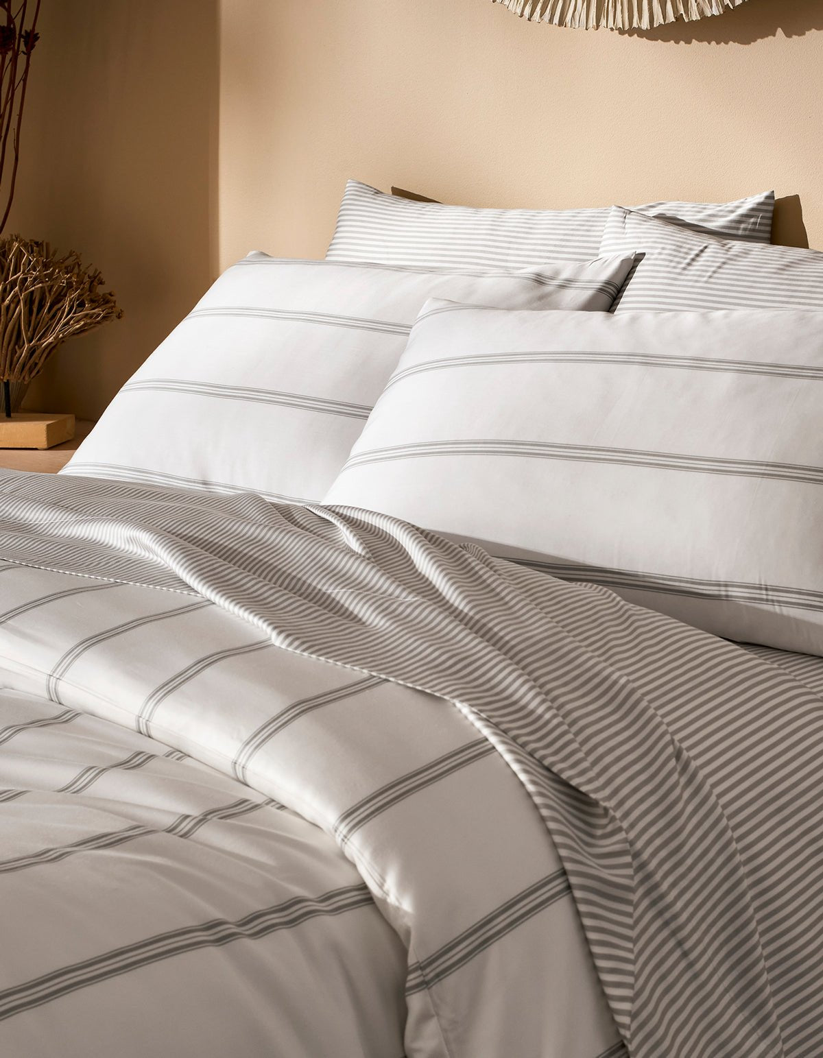 A neatly made bed features the Cozy Earth Bamboo Duvet Cover in white with light gray stripes, two large pillows, and a cozy comforter. The neutral background includes a nightstand with decorative dried plants.|Color:Baja Stripe in Laurel